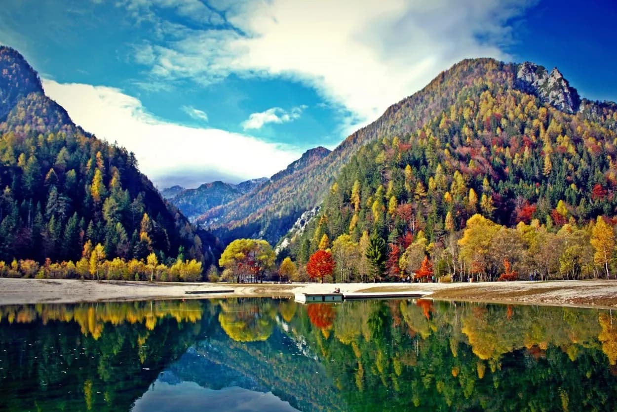Alpine lake reflecting mountains with vibrant autumn foliage under a blue sky at Kranjska Gora Jezero Jasna.