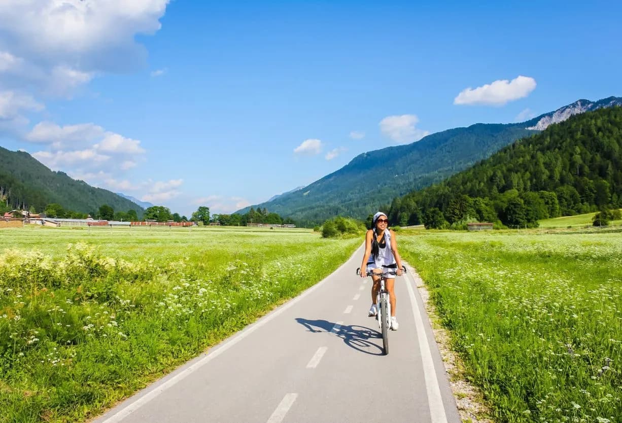 Woman cycling on path through green meadow with forested Alps mountains in background