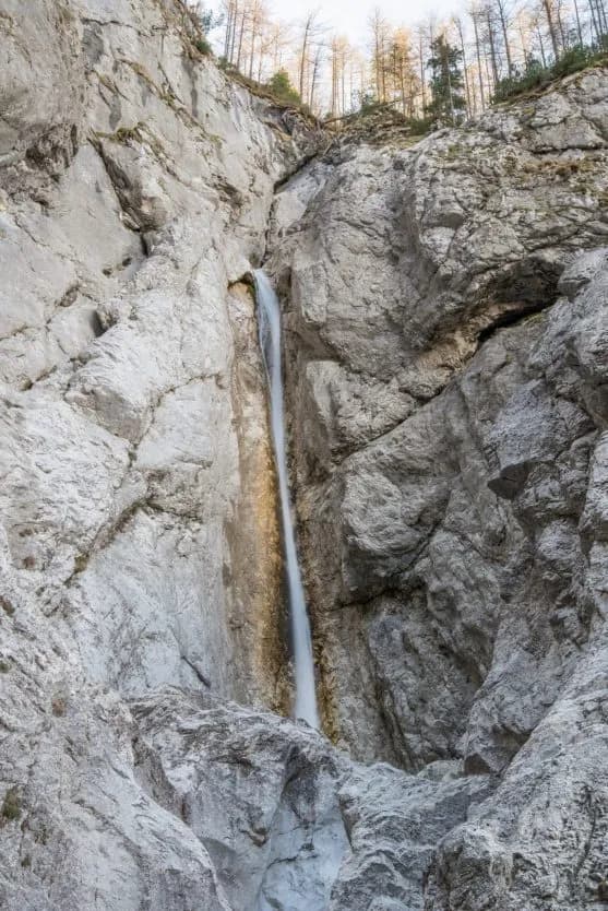 Tall, narrow waterfall cascading down steep, gray rock cliffs with sparse trees at the top.