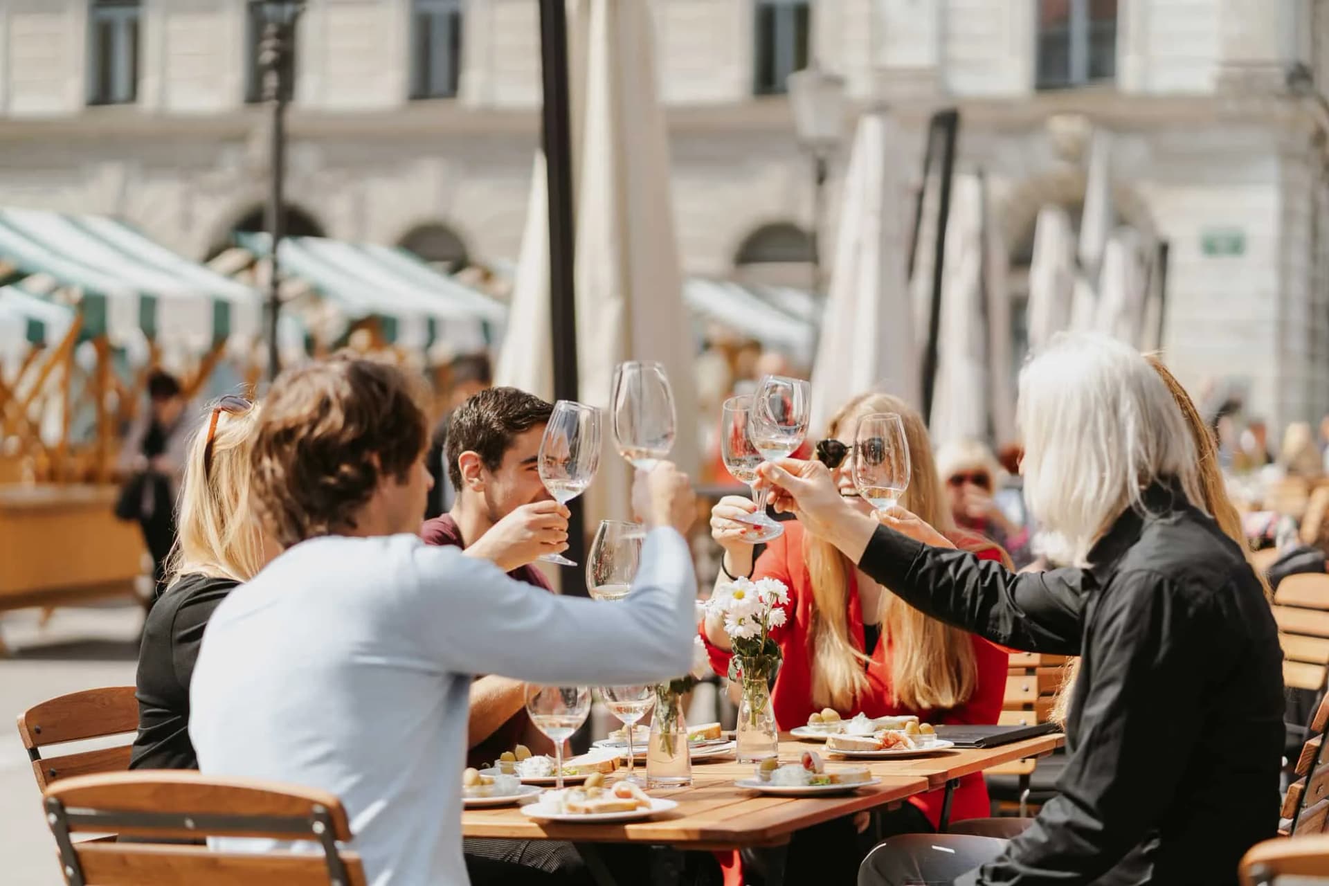 Group toasting with wine glasses at outdoor cafe table with food and flowers.