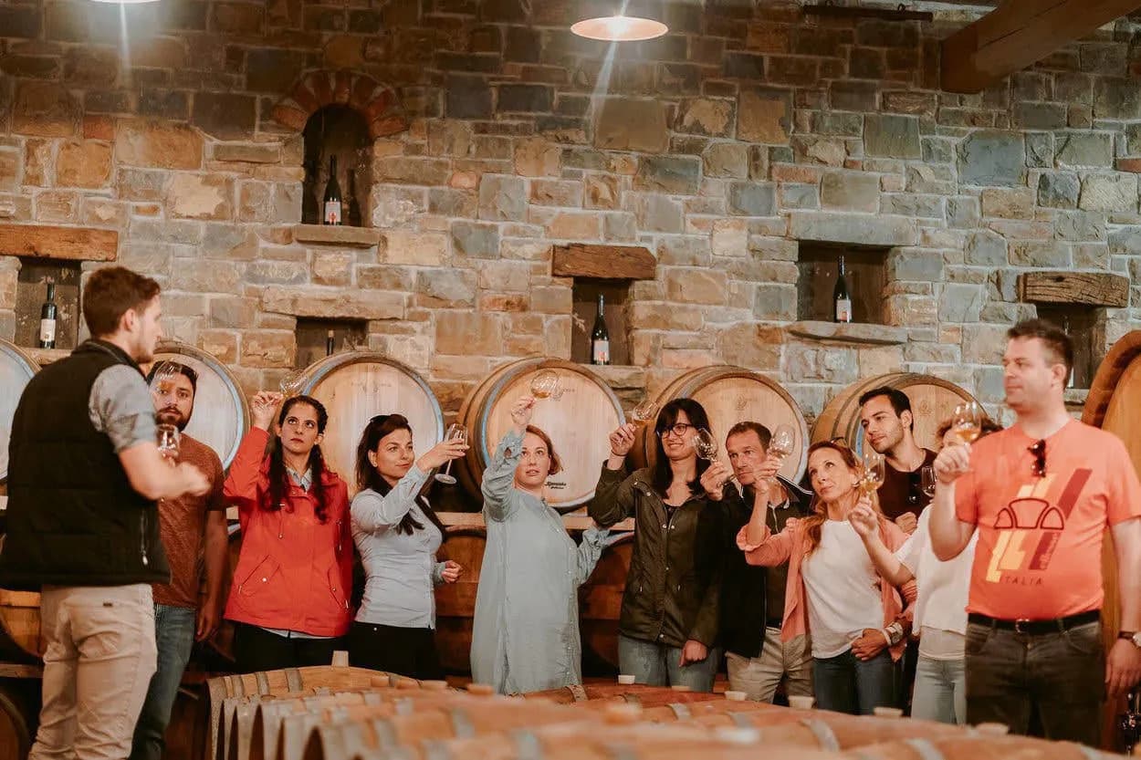 Wine tasting tour group holding glasses in a cellar with large wooden barrels and stone walls.