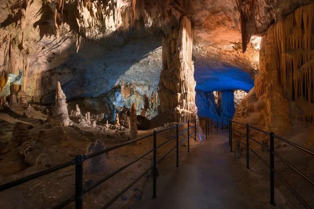 Postojna Cave interior with illuminated stalactites, stalagmites, and a visitor pathway.