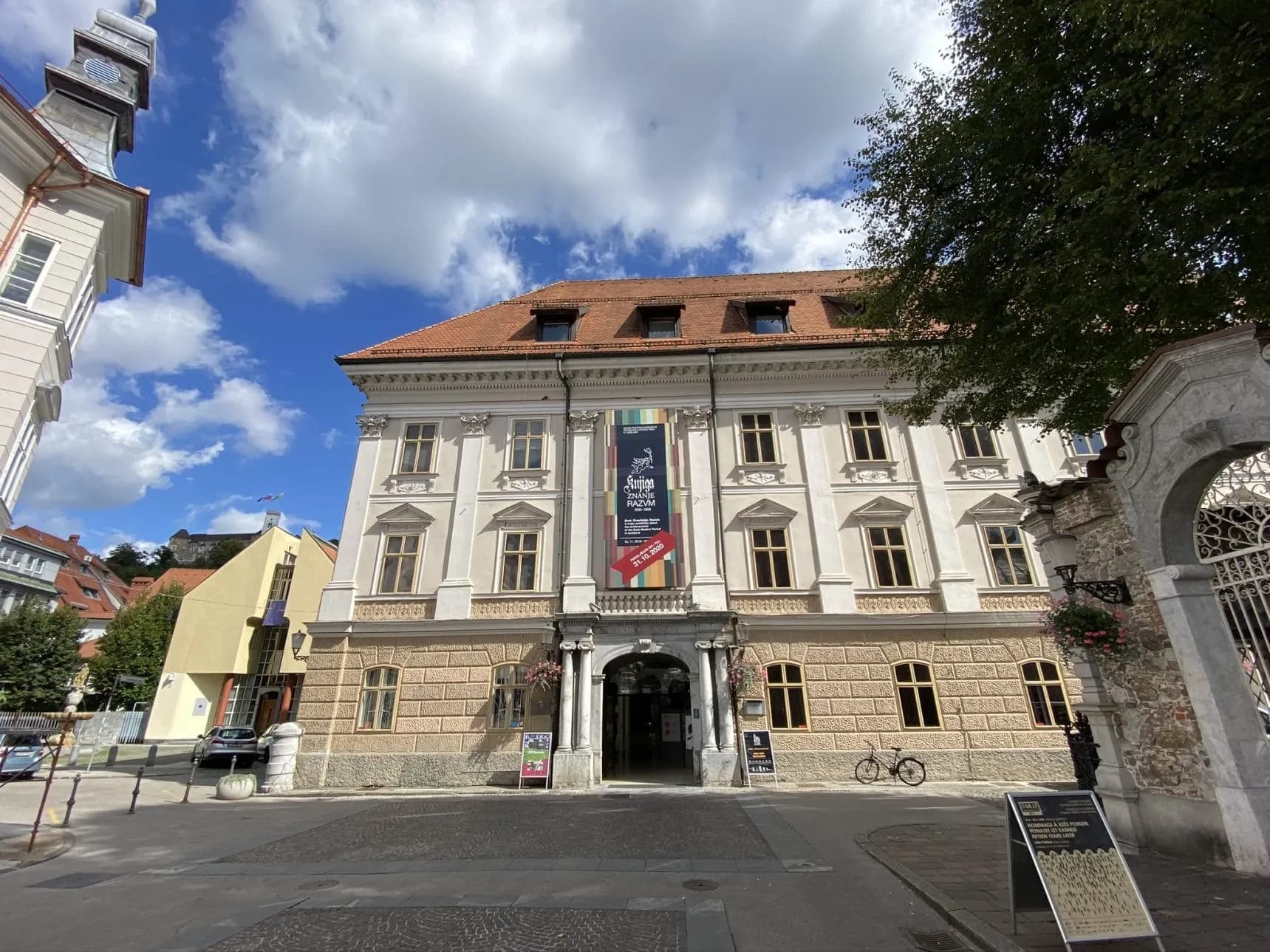 Ljubljana City Museum building facade with banner under a bright, cloudy sky.