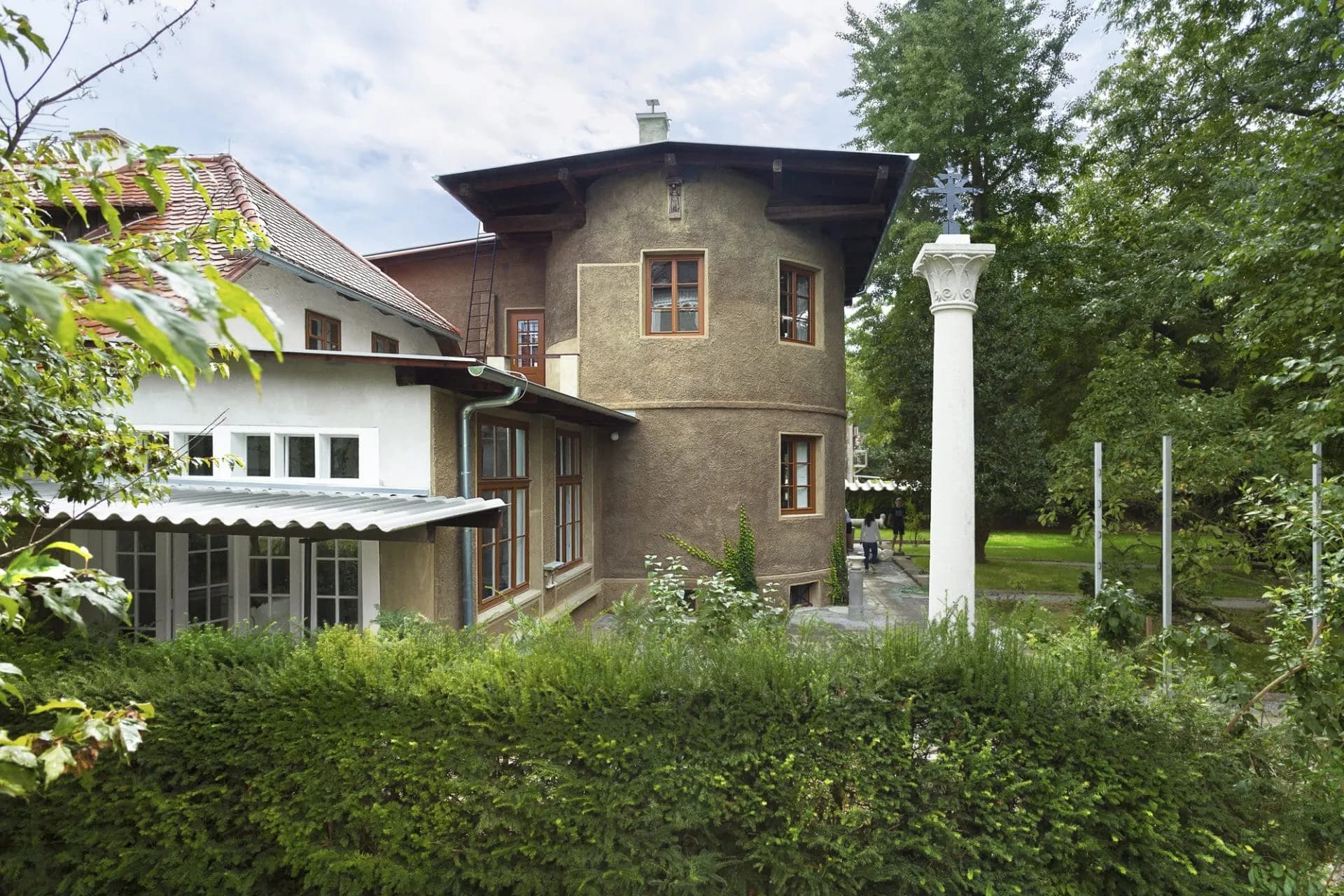 Plečnik House with stucco facade, prominent round tower, and white column topped with a cross.