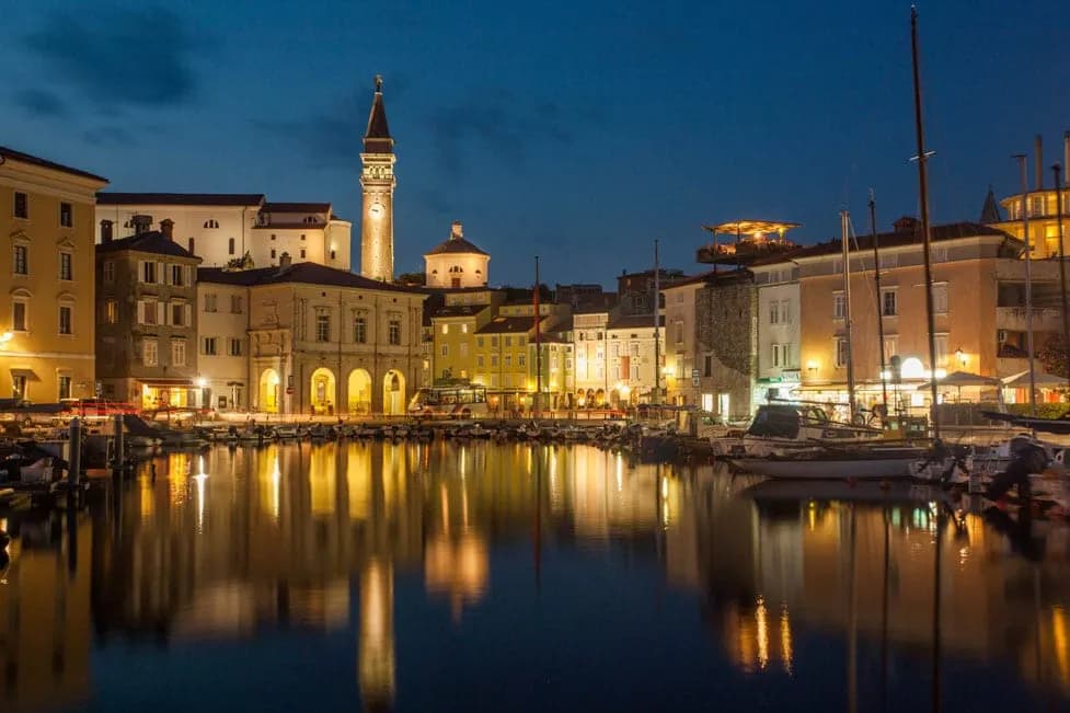 Boats docked in harbor reflecting illuminated buildings and clock tower at night in Piran.