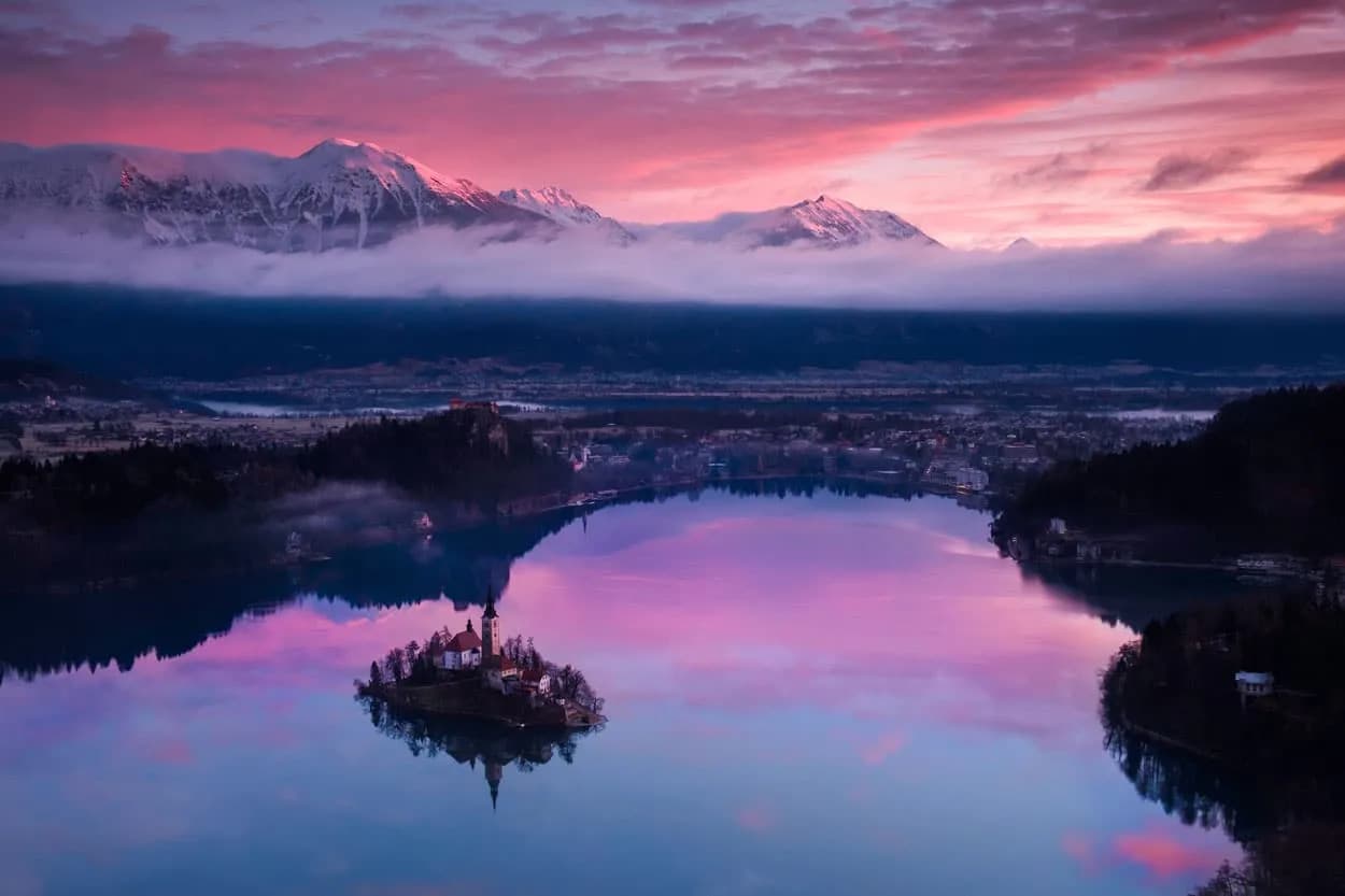 Lake Bled island church reflected in water at sunrise with snow-capped mountains.