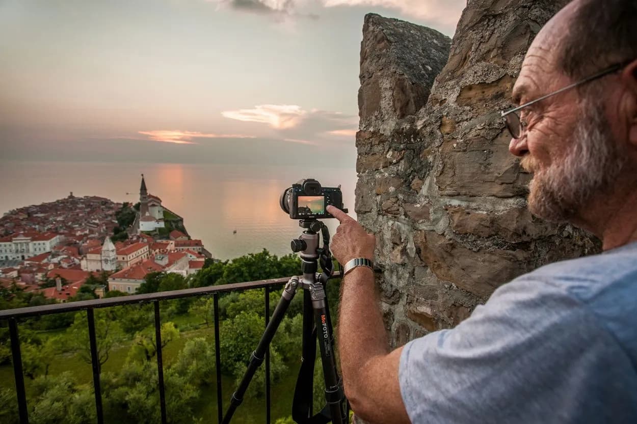 Photographer adjusting camera on tripod overlooking Piran, Slovenia coastal town at sunset.