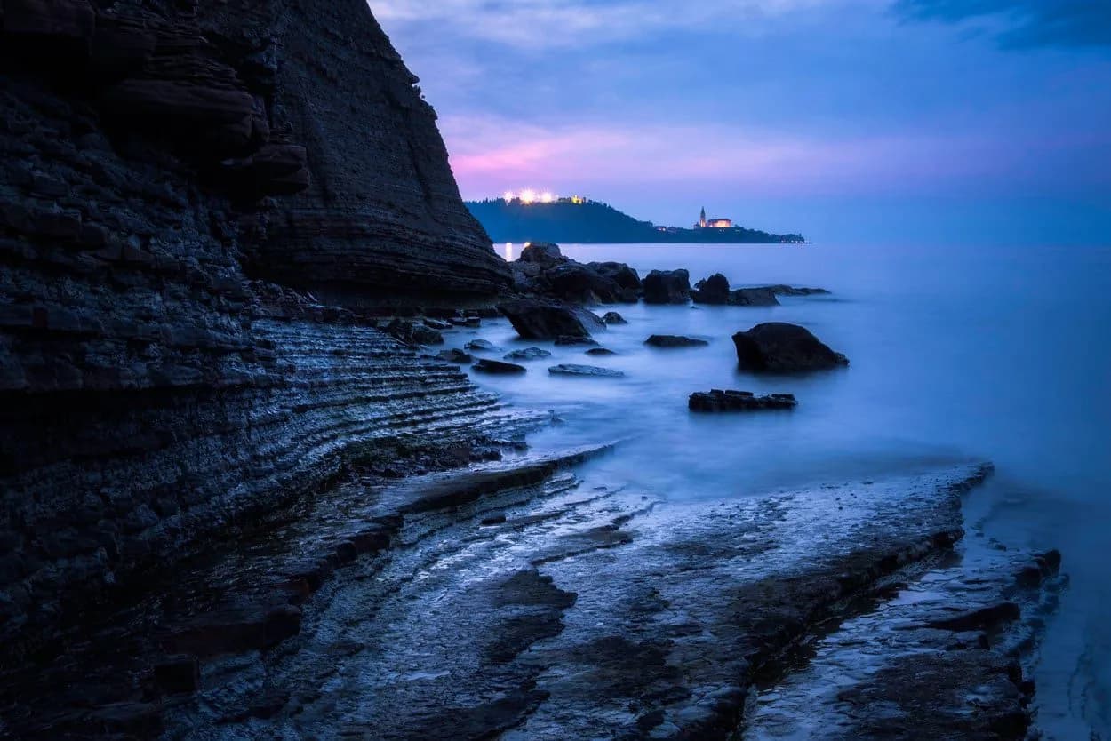 Layered coastal rocks at dusk with calm sea and distant illuminated town in Slovenia.