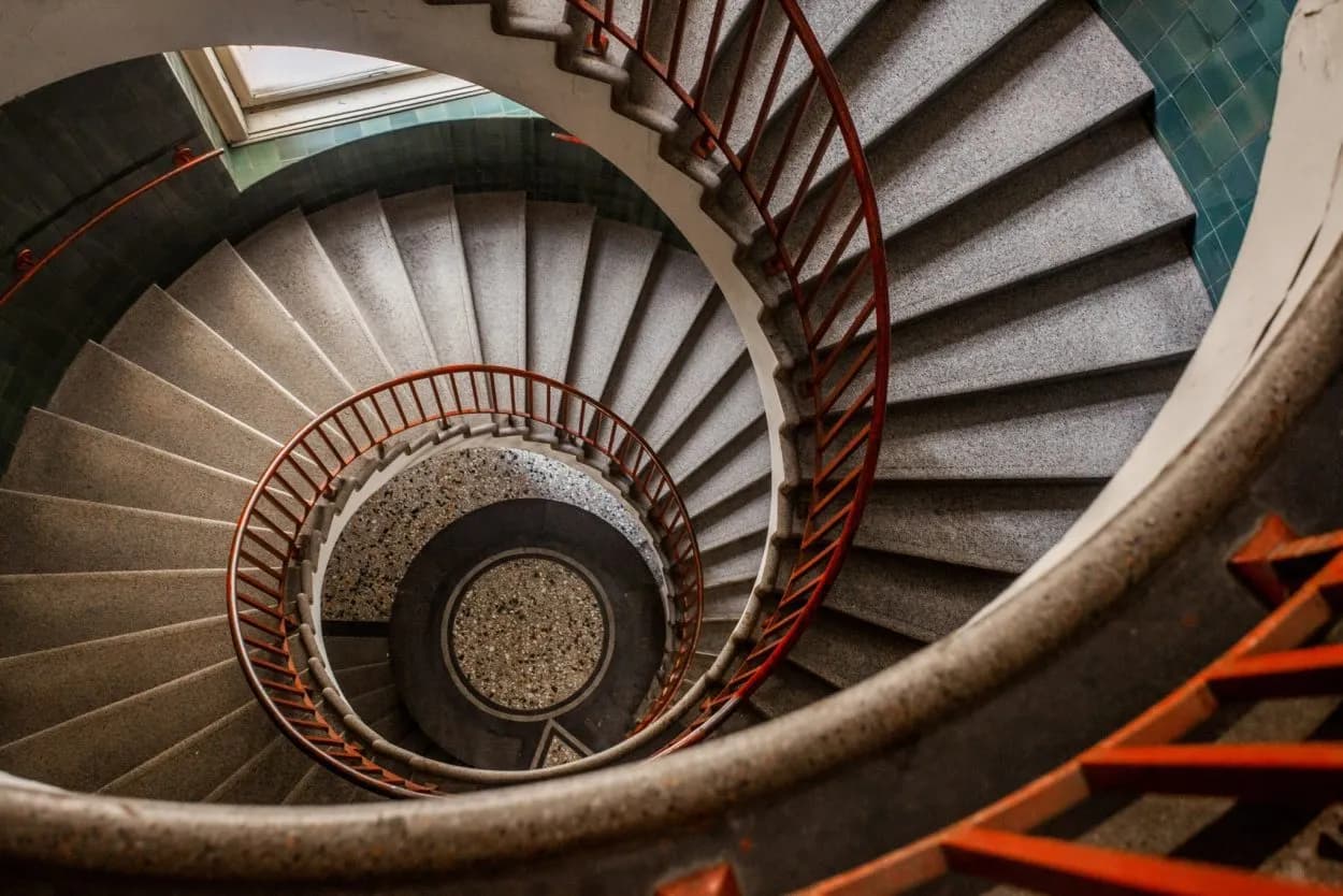 Spiral staircase with grey stone steps and red metal railing descending into a central well in Ljubljana.