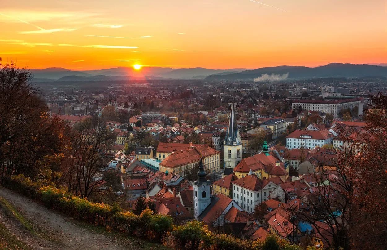 Cityscape of Ljubljana with church spires at sunset overlooking mountains, viewed from a hillside.