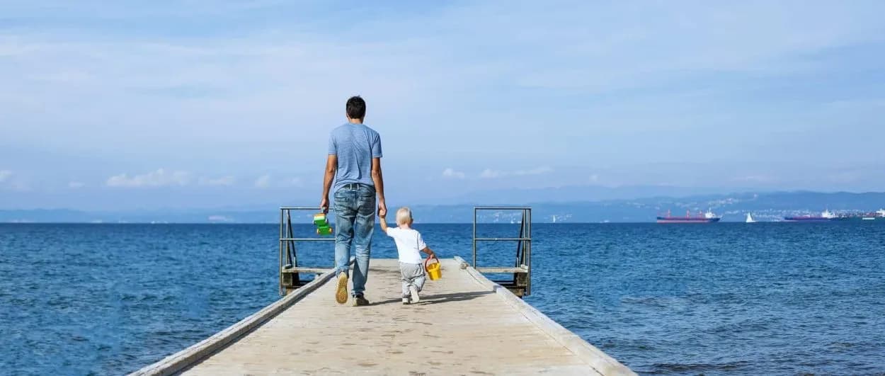 Father and child walking on pier toward blue sea with distant cargo ships and mountains.
