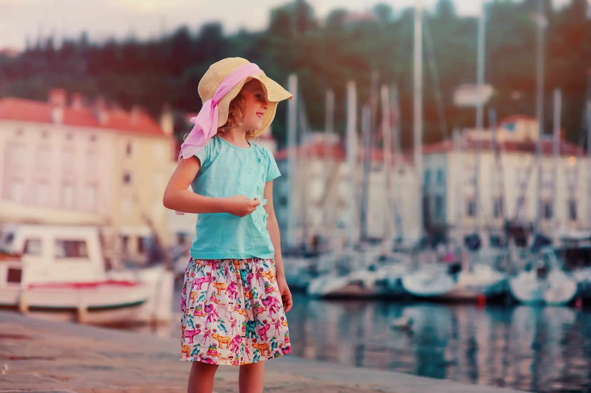 Girl in sun hat standing by marina with docked sailboats and coastal buildings