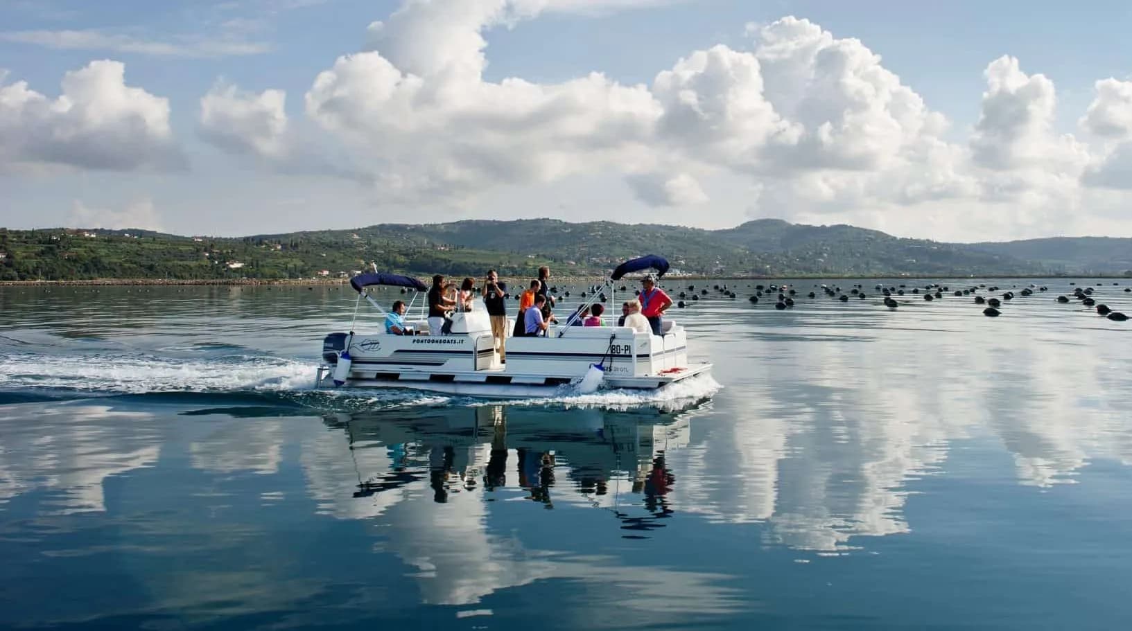 Pontoon boat carrying passengers on calm water near coastal hills and aquaculture buoys