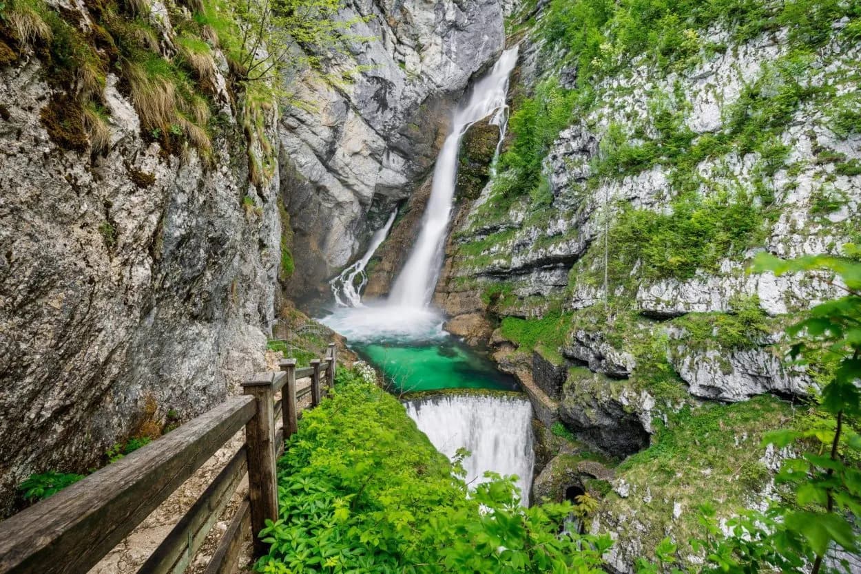Waterfall cascading into a turquoise pool beside a wooden path in a lush, rocky gorge.