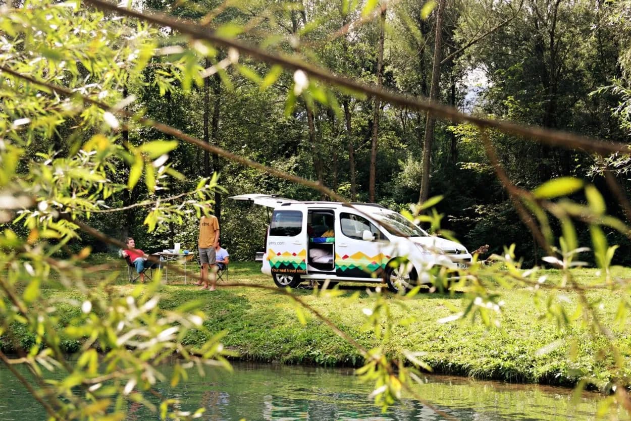Camper van parked by a river with people relaxing in camping chairs in a forest setting.