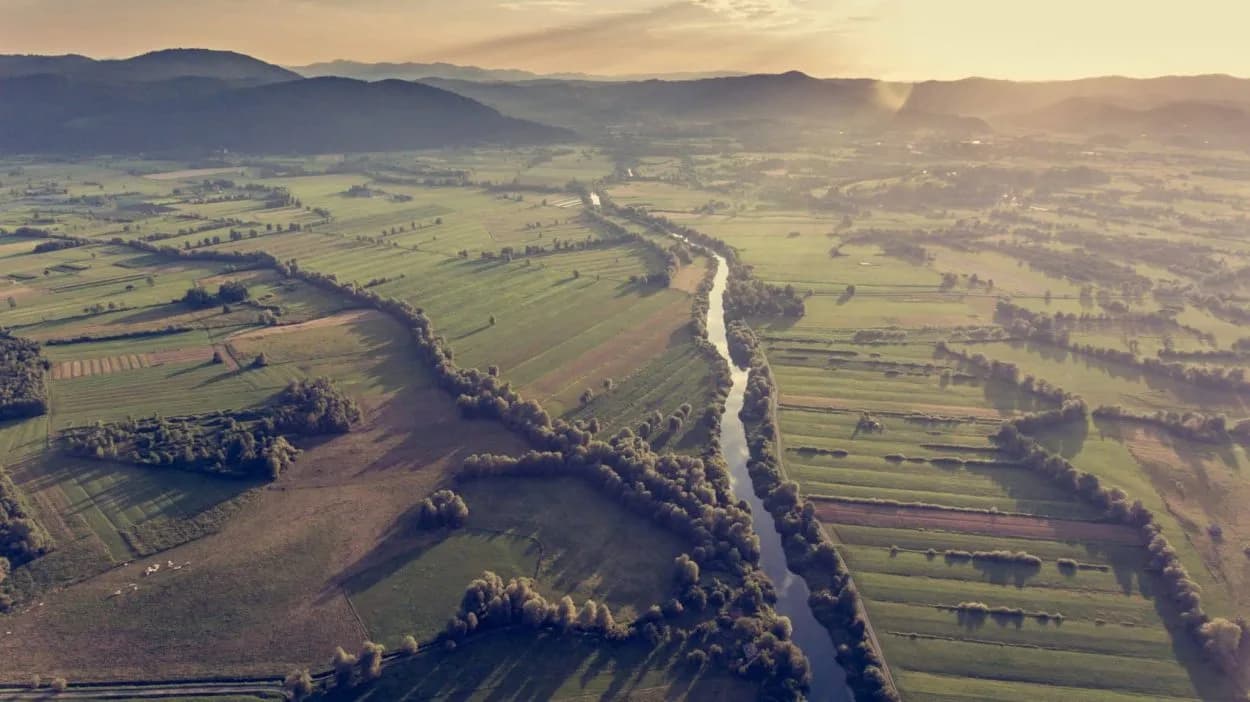 Aerial view of river winding through green patchwork fields with rolling mountains at sunset
