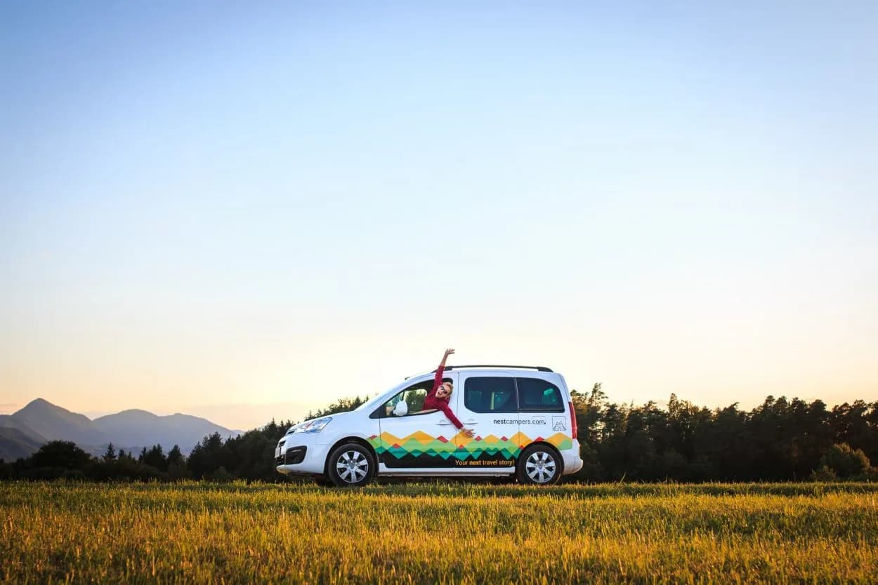 Camper van parked in grassy field with mountains and forest at sunset
