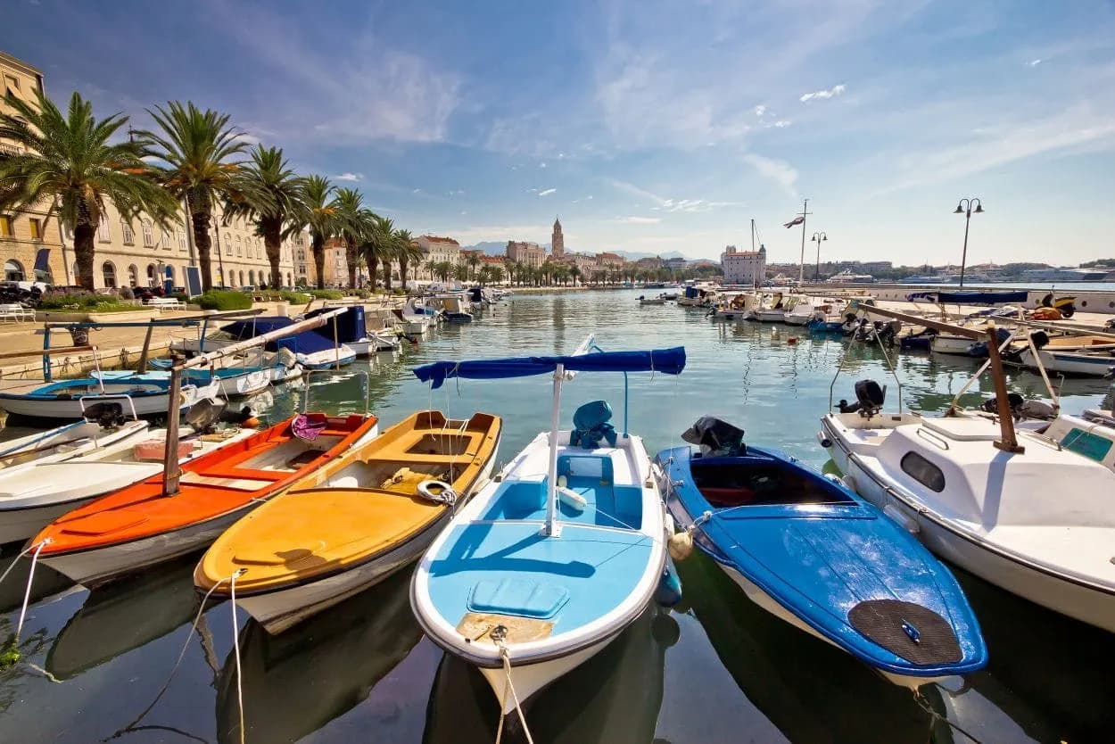 Colorful small boats moored in a harbor with palm trees and historic buildings in the background.