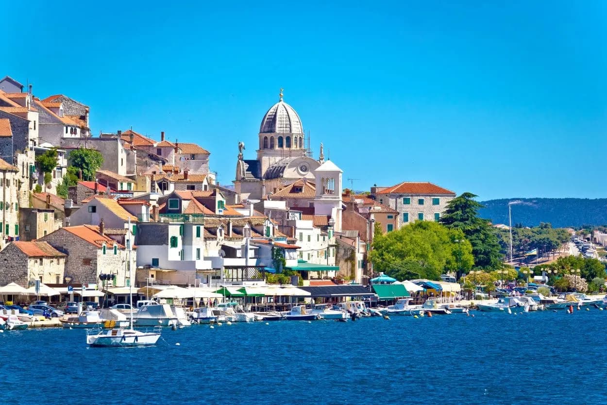 Sibenik waterfront with historic buildings, cathedral dome, and boats on blue sea.