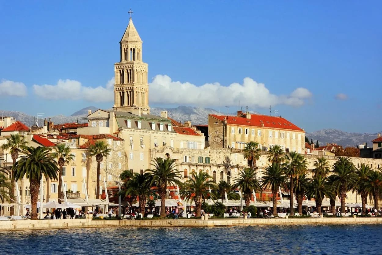 Bell tower overlooking waterfront cafes, palm trees, and mountains in Split, Croatia