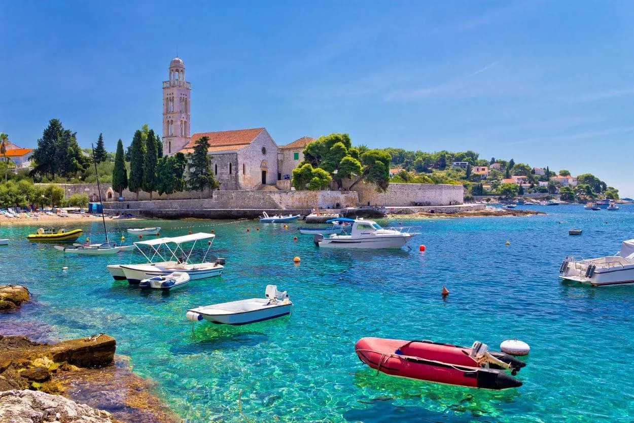 Boats in turquoise water near stone church with bell tower on Hvar island.