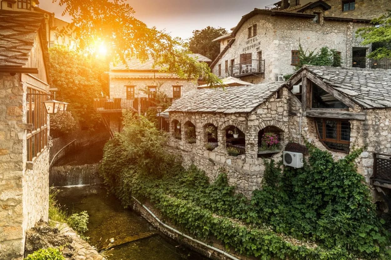Stone buildings with slate roofs beside a narrow river with a small waterfall in Mostar old town.