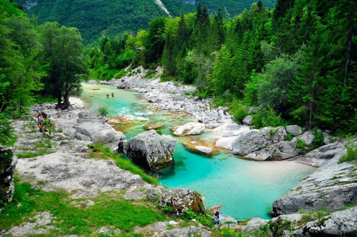Hikers near turquoise river with clear pools surrounded by lush green forest mountains.