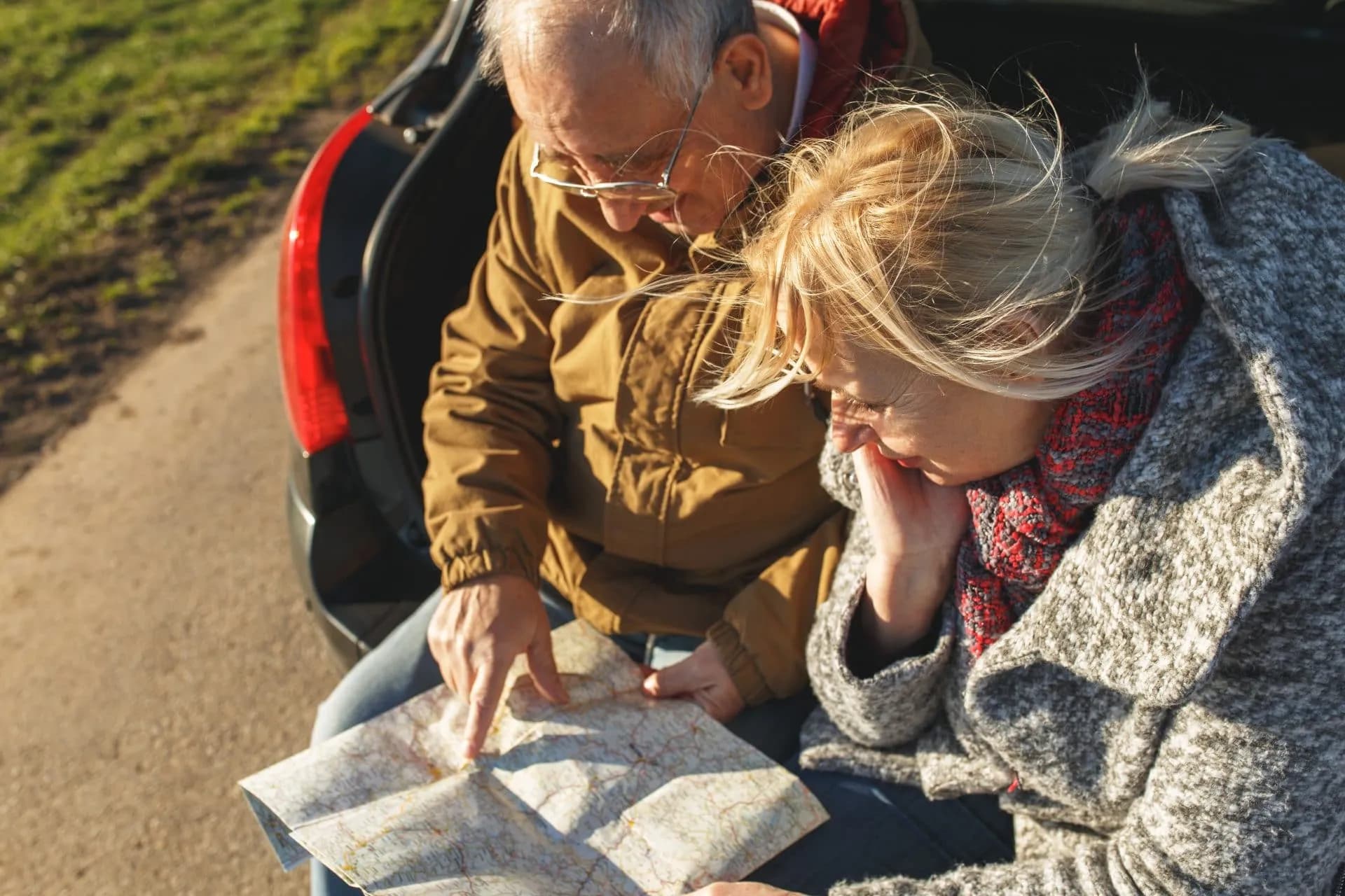 Senior couple taking a break, looking at a paper map next to their car on a roadside.