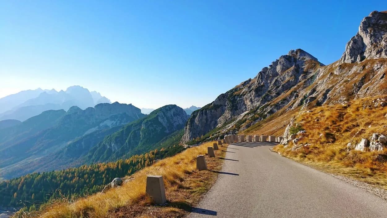 Mountain road winding past rocky slopes with autumn grass and layered peaks in Slovenia.
