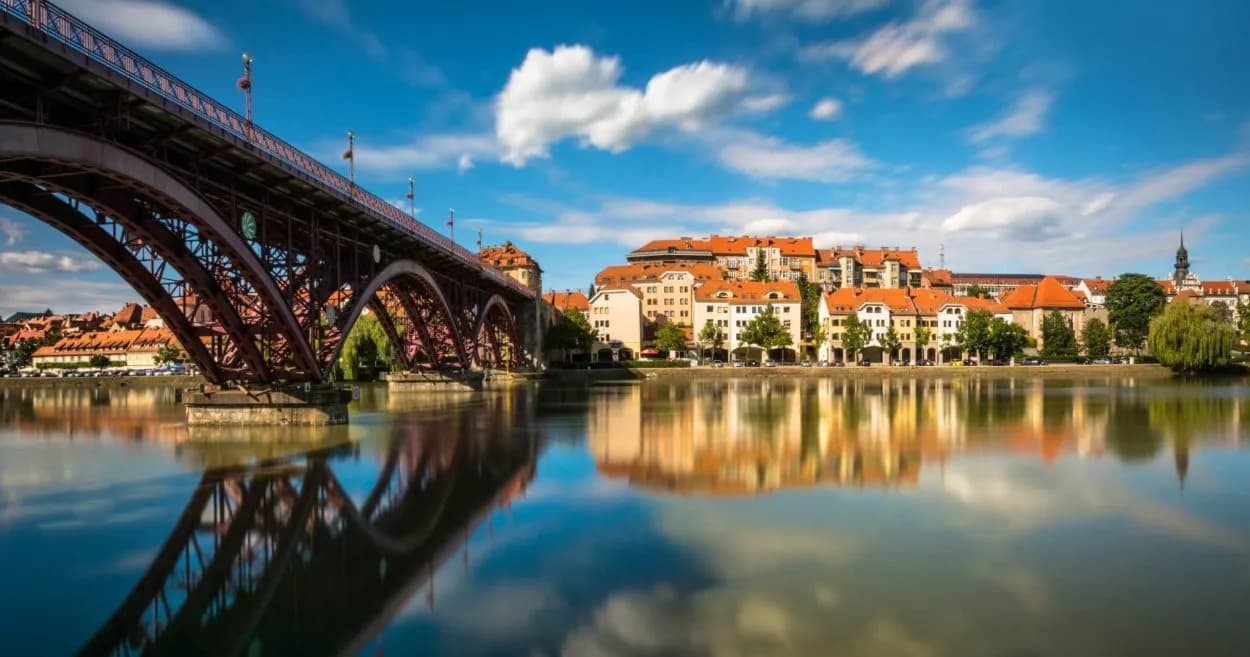 Maribor bridge over river reflecting terracotta-roofed buildings under blue sky