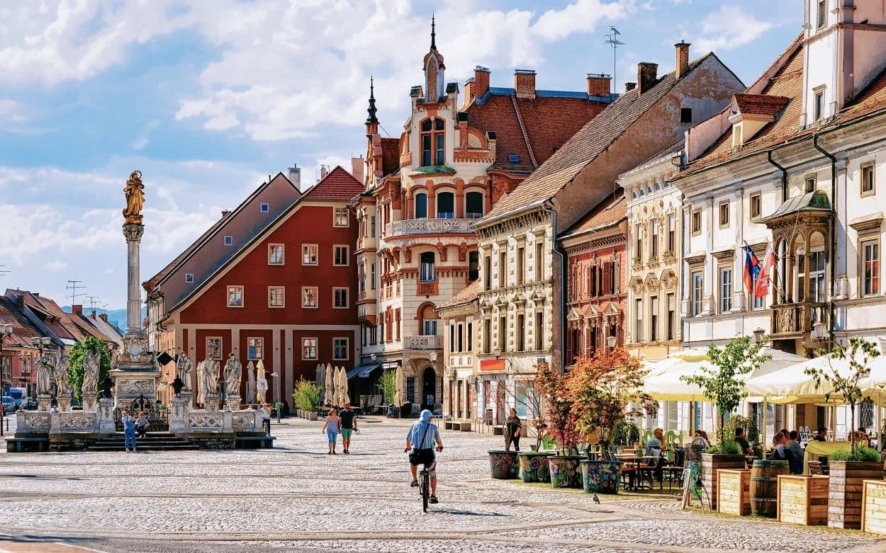 Cobblestone square in Maribor with plague column, historic buildings, and outdoor cafe seating.
