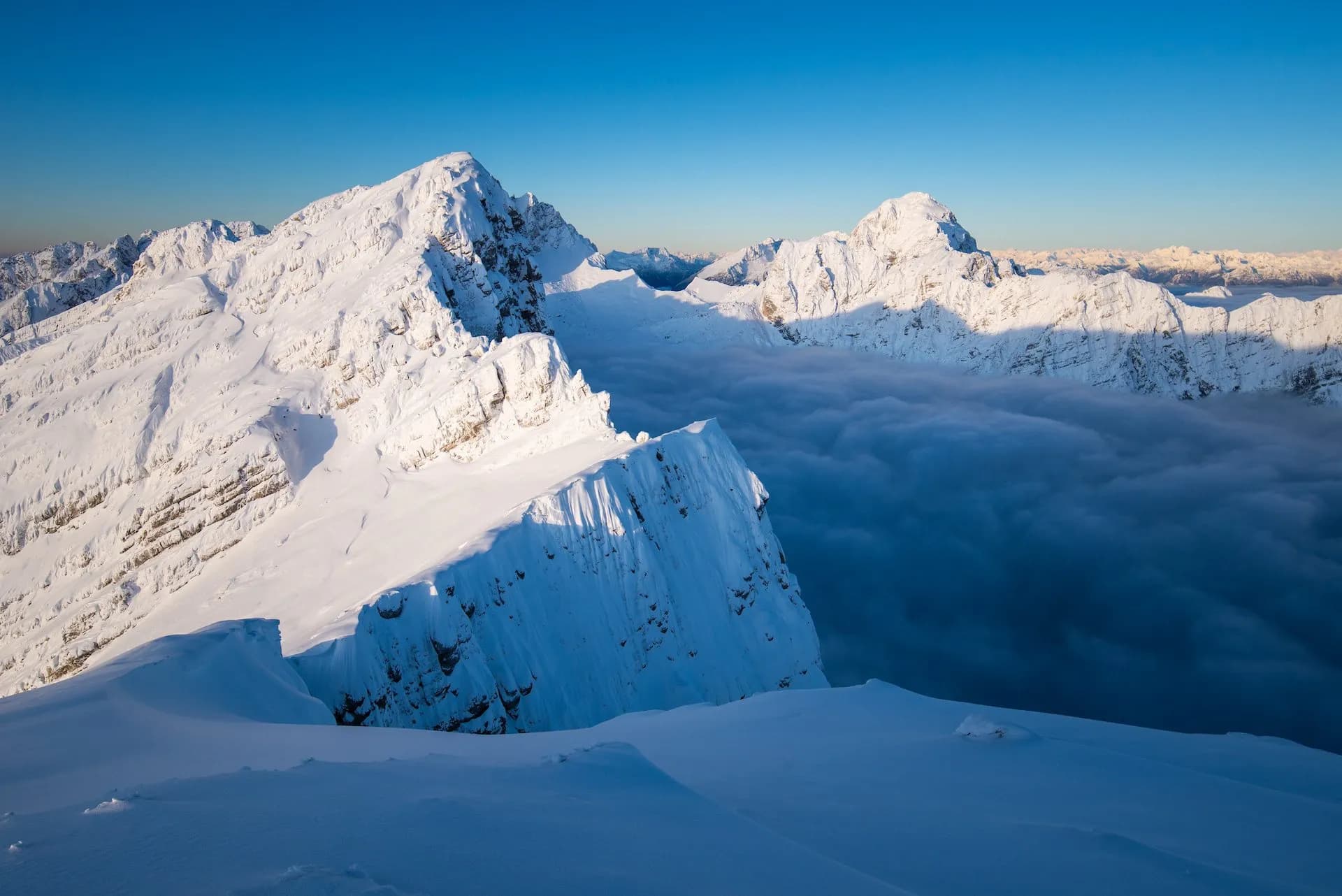 Snowy mountain peaks above a sea of clouds under a clear blue sky from Mojstrovka.