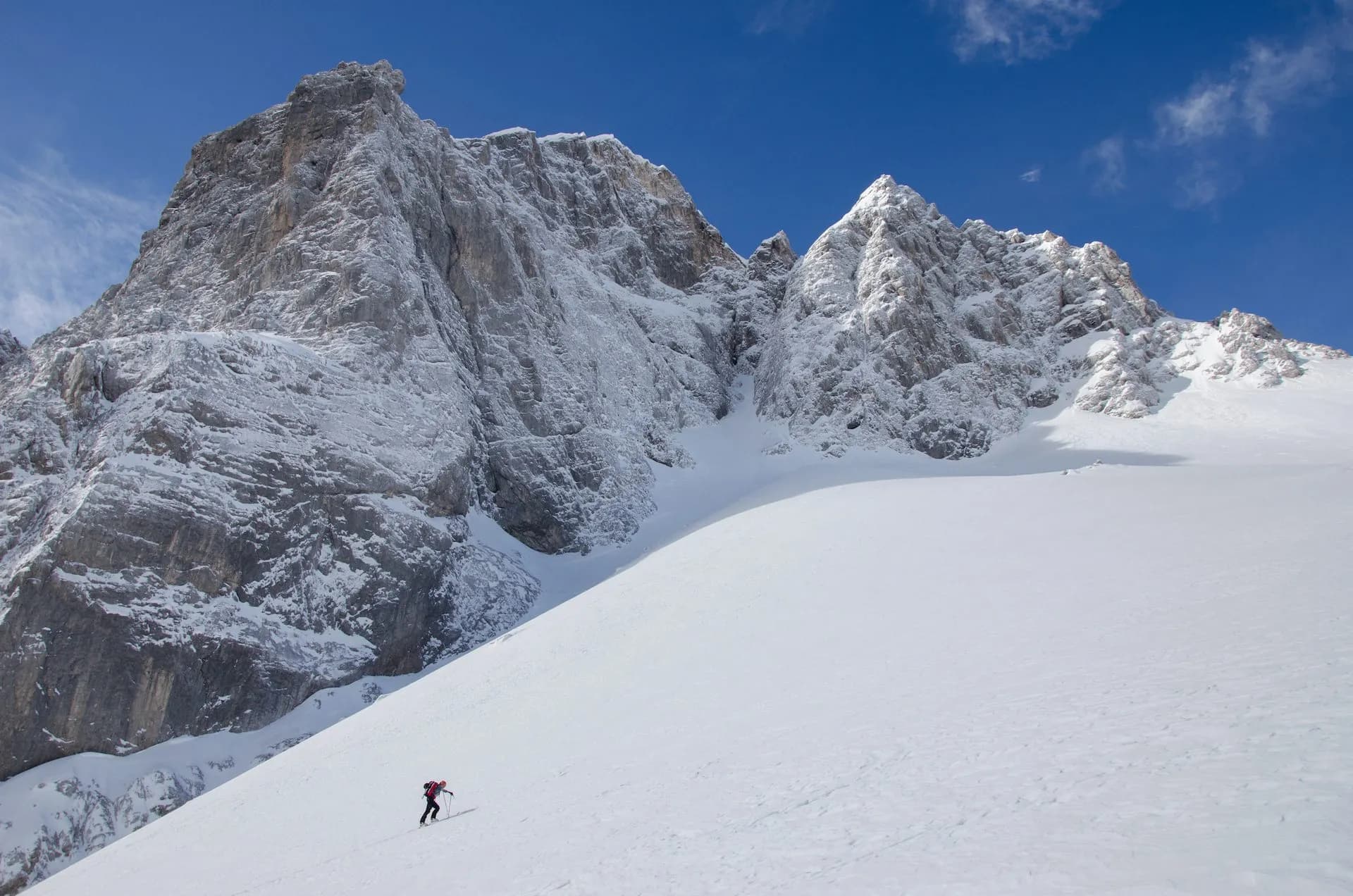 Ski touring up a vast snowfield toward massive, snow-dusted rocky mountains under a blue sky.
