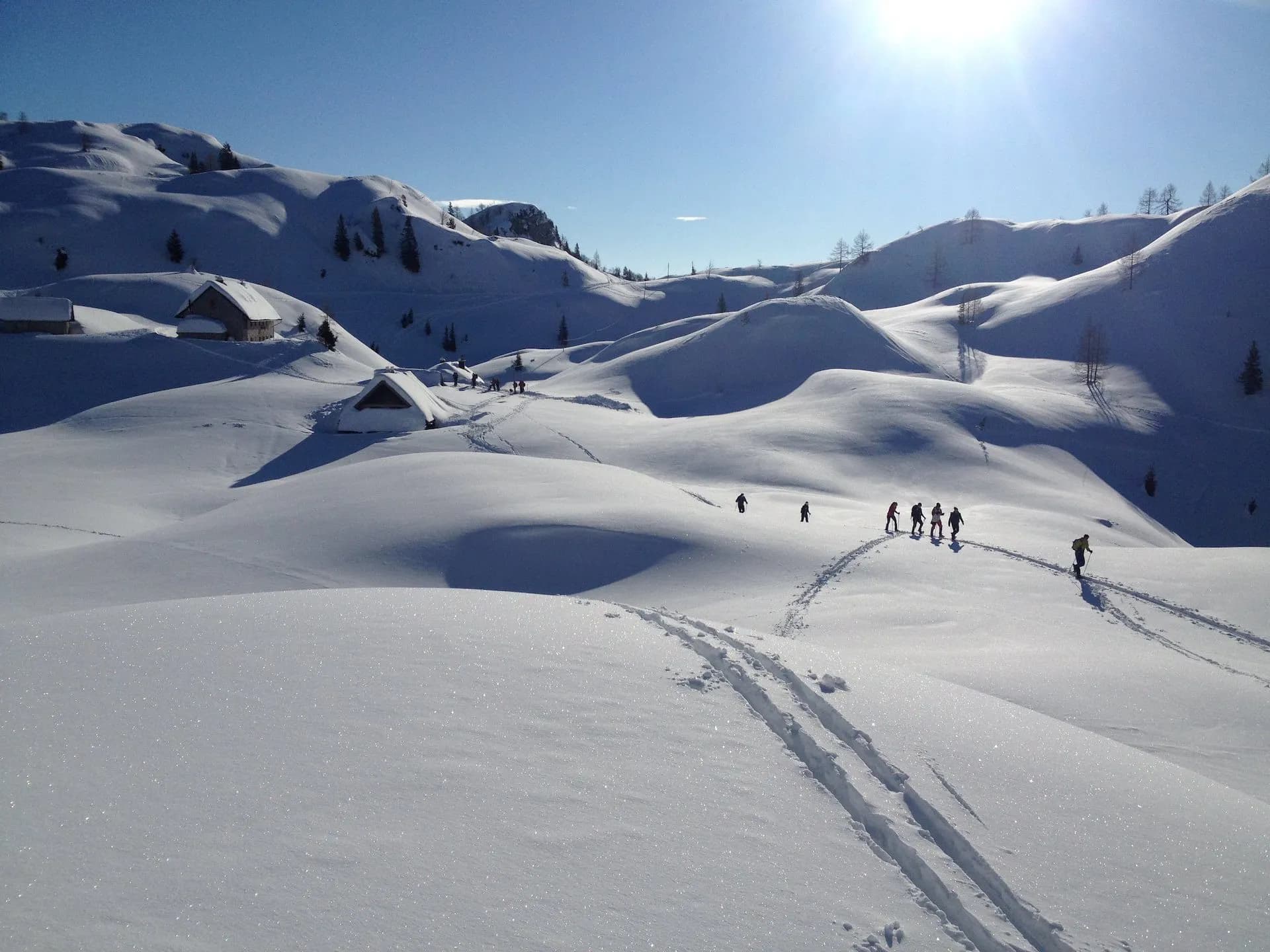 Hikers traversing deep snow on Komna Plateau with small wooden huts visible in the background.