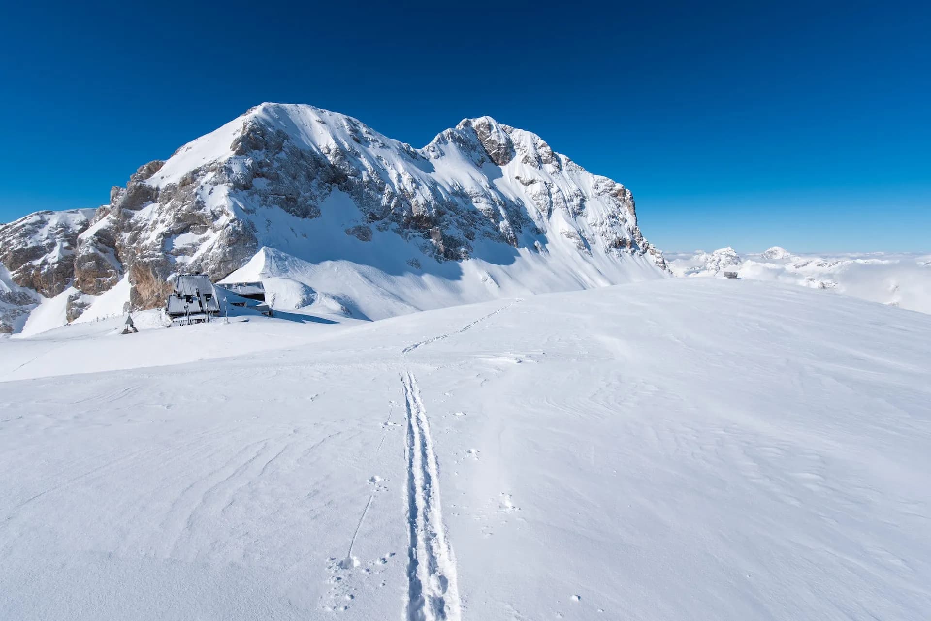 Ski tracks lead toward Kredarica and Triglav mountains in deep winter snow under a clear blue sky.