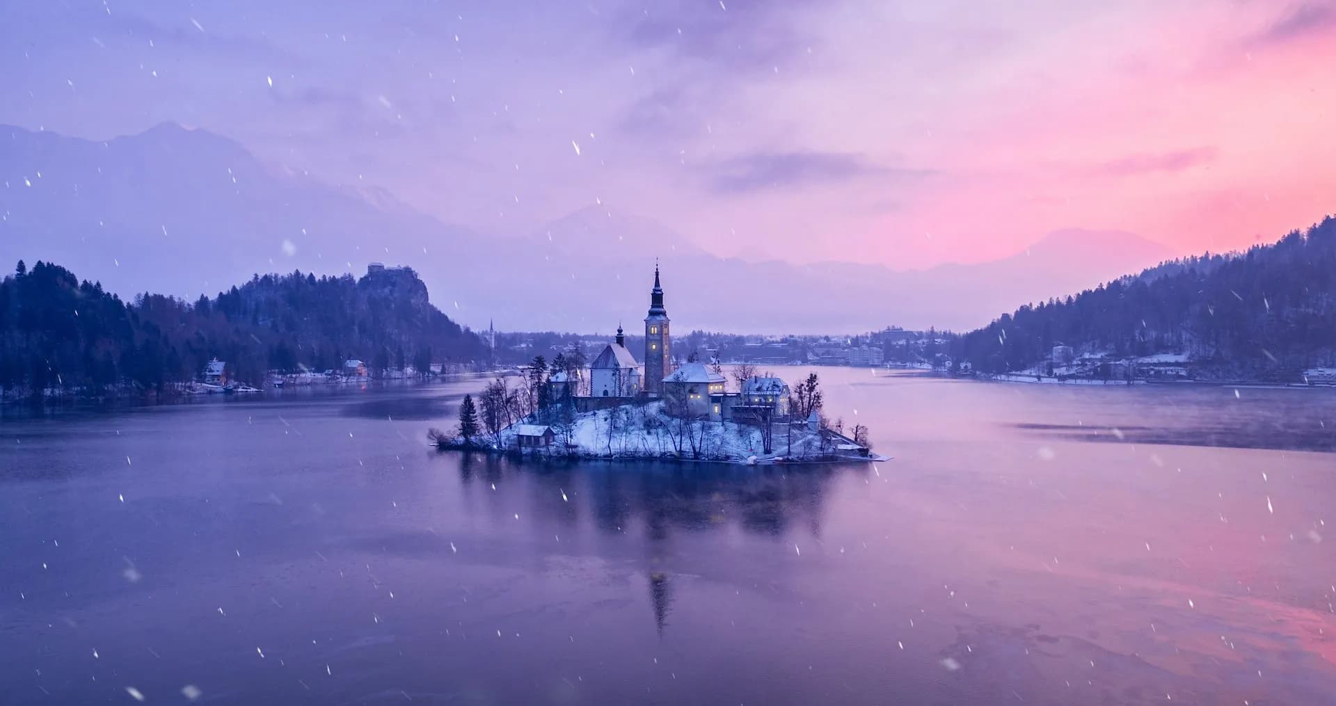 Church on snow-covered island in Lake Bled during winter sunset with falling snow