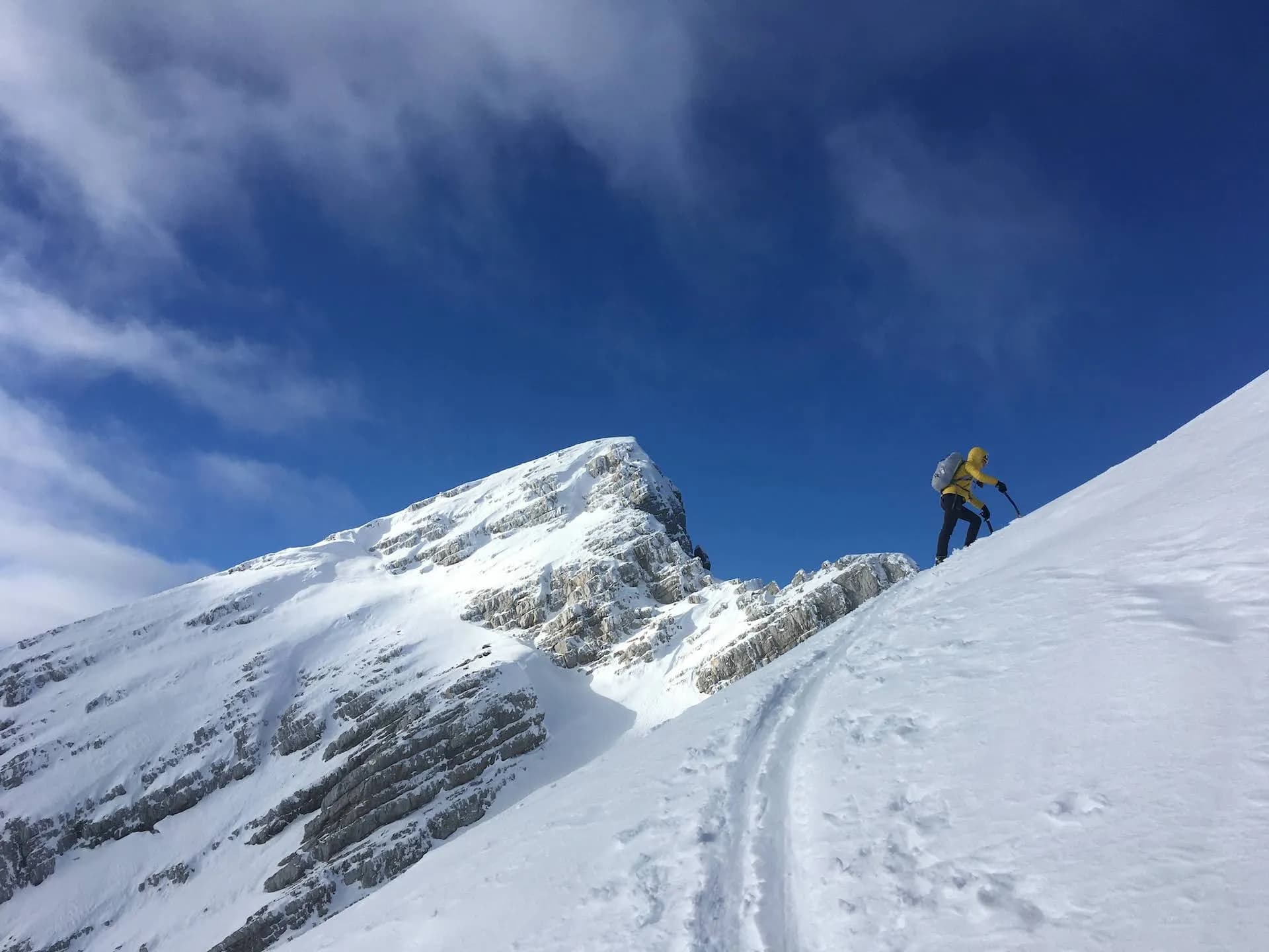 Ski mountaineer ascending steep snowy slope below Mala Mojstrovka peak under blue sky.