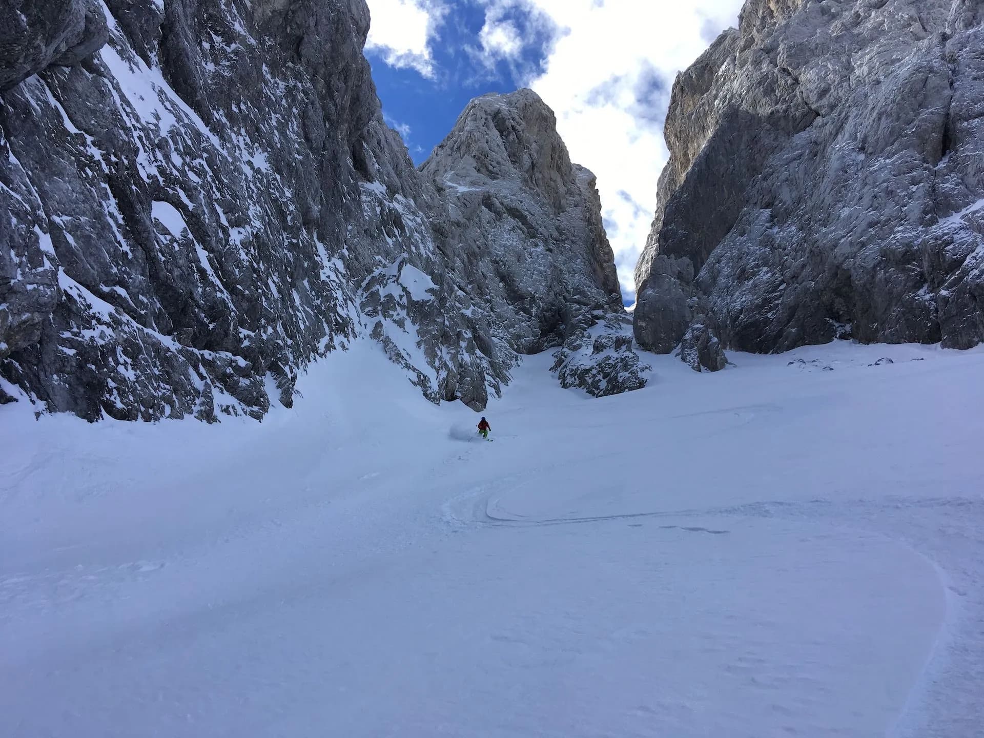 Skier descending deep powder snow between steep, rocky, snow-covered mountains under blue sky.