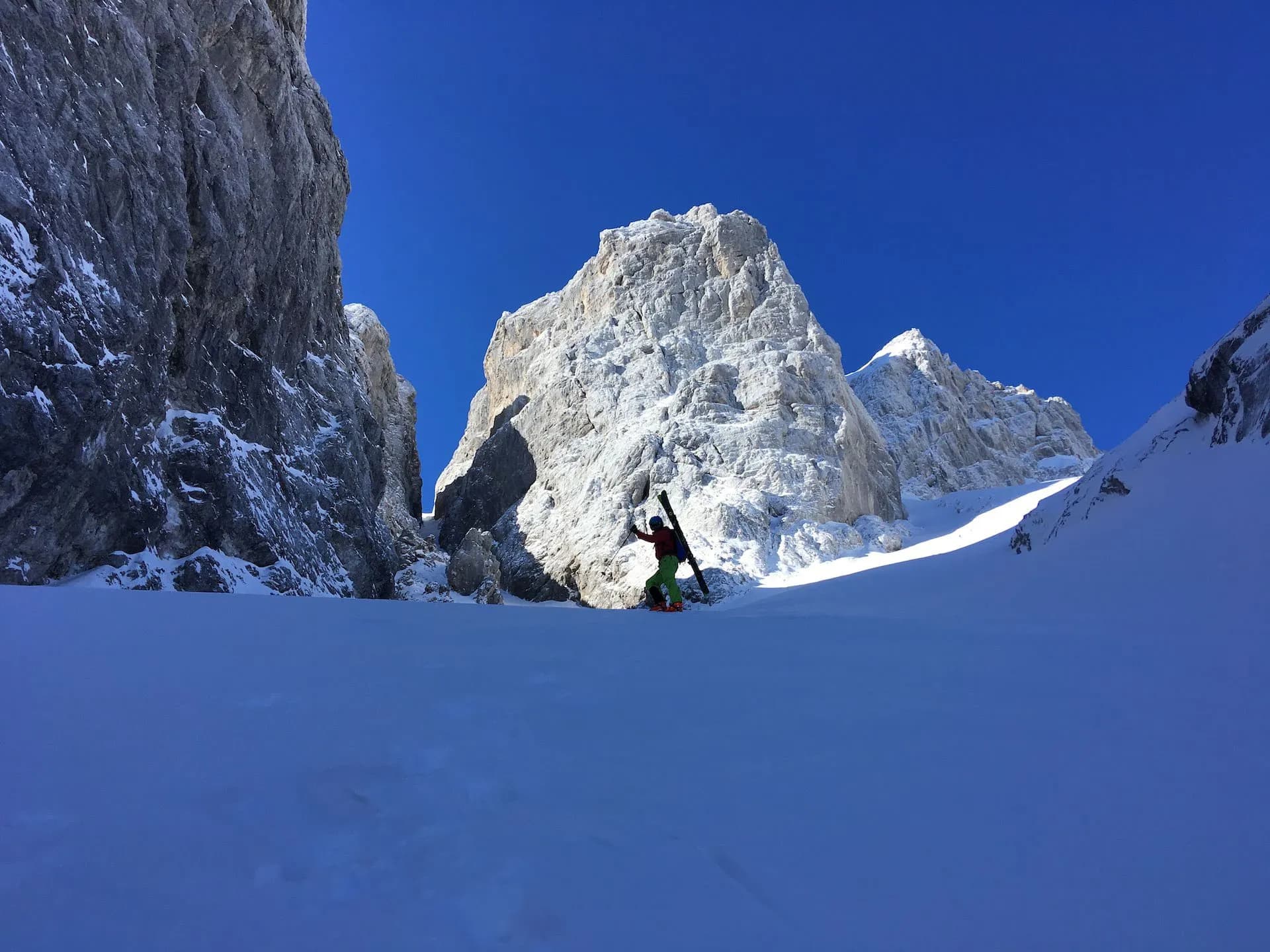 Ski mountaineer hiking up deep snow below massive, sunlit, rocky peaks under a clear blue sky.