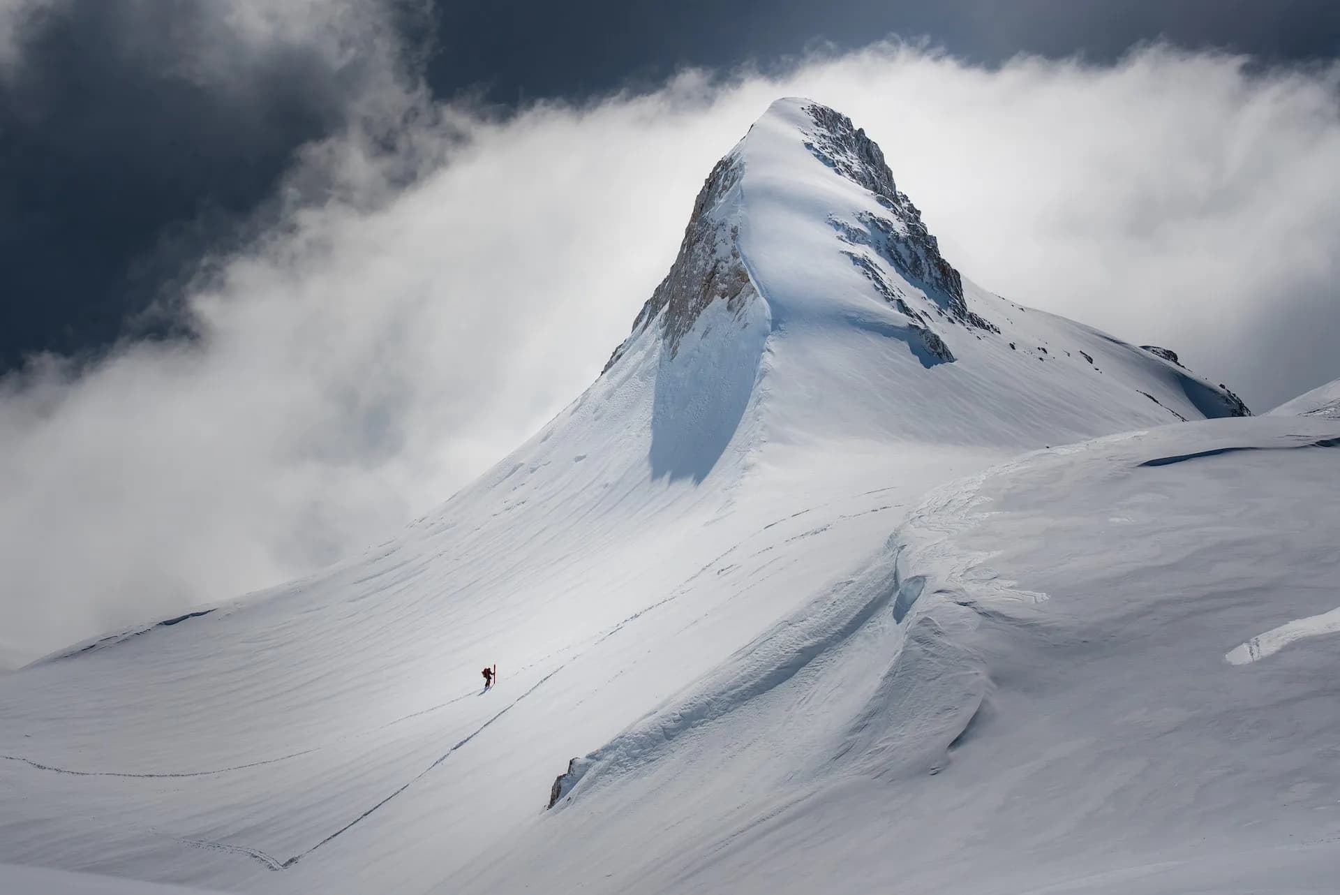 Ski touring below Kredarica on a vast, snow-covered mountain slope under dramatic clouds.