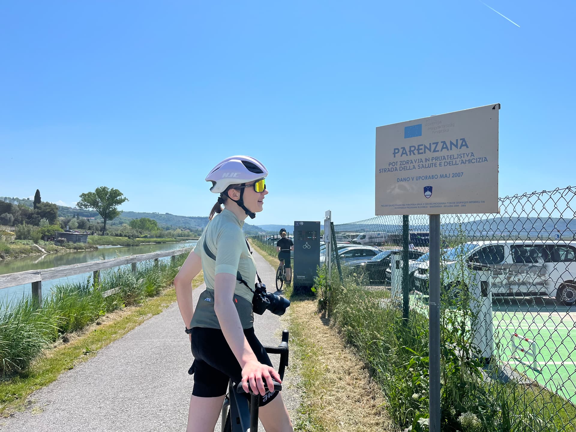 Cyclist with camera stops near Parenzana sign beside waterway and parking lot.