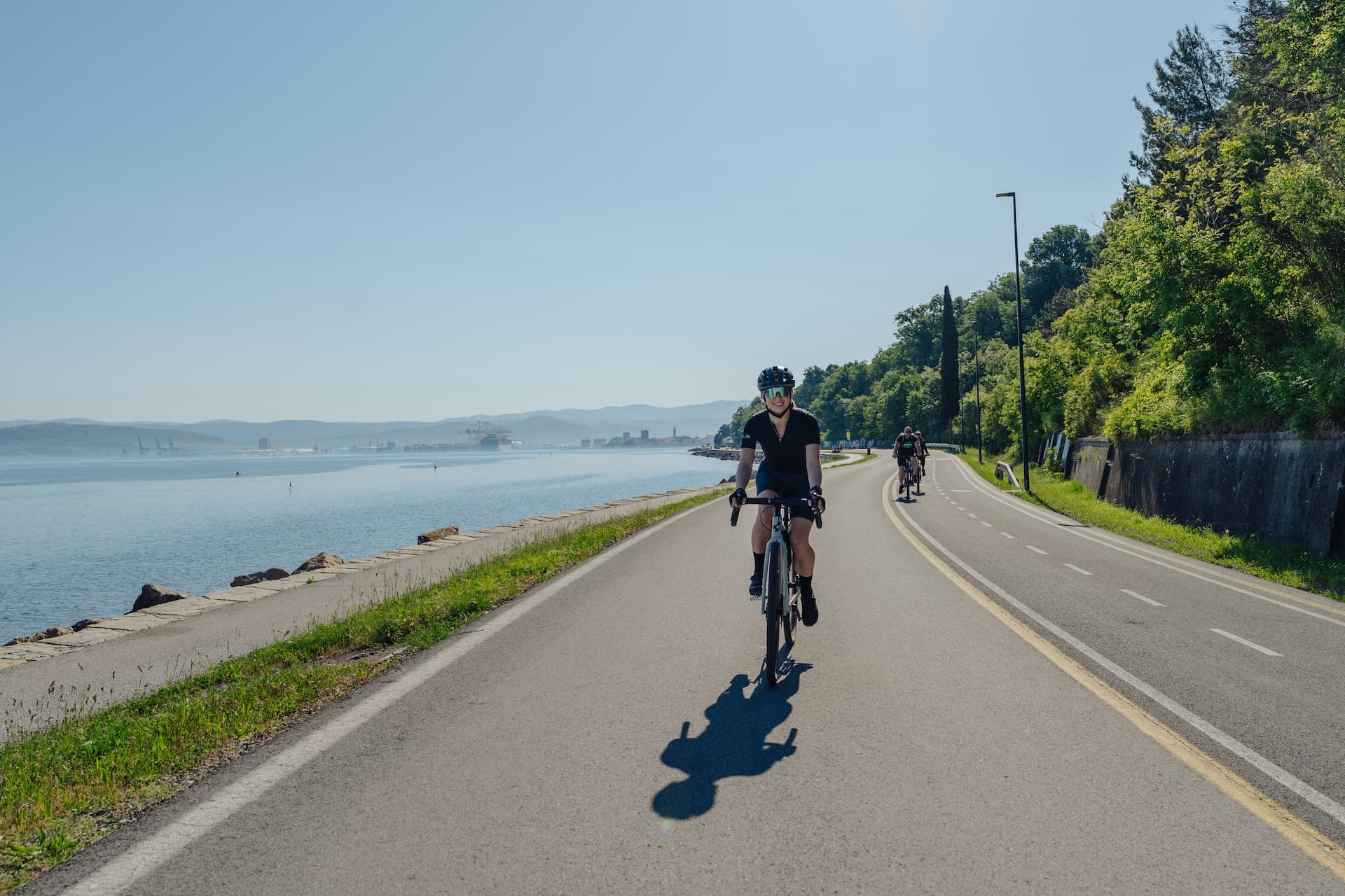 Cyclists riding road bikes along a sunny coastal road next to the sea.