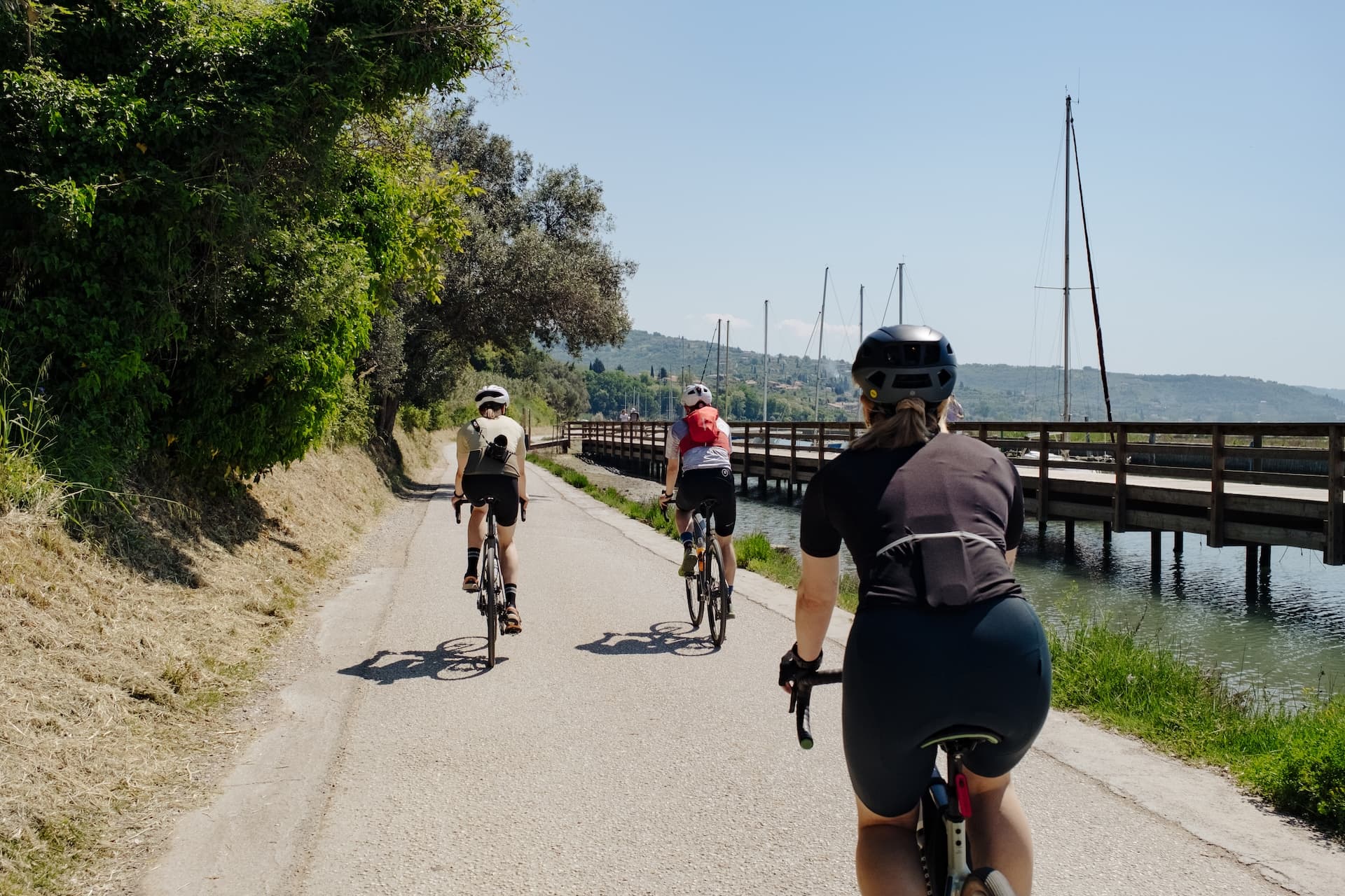 Cyclists riding on paved path near harbor with sailboats and green hills