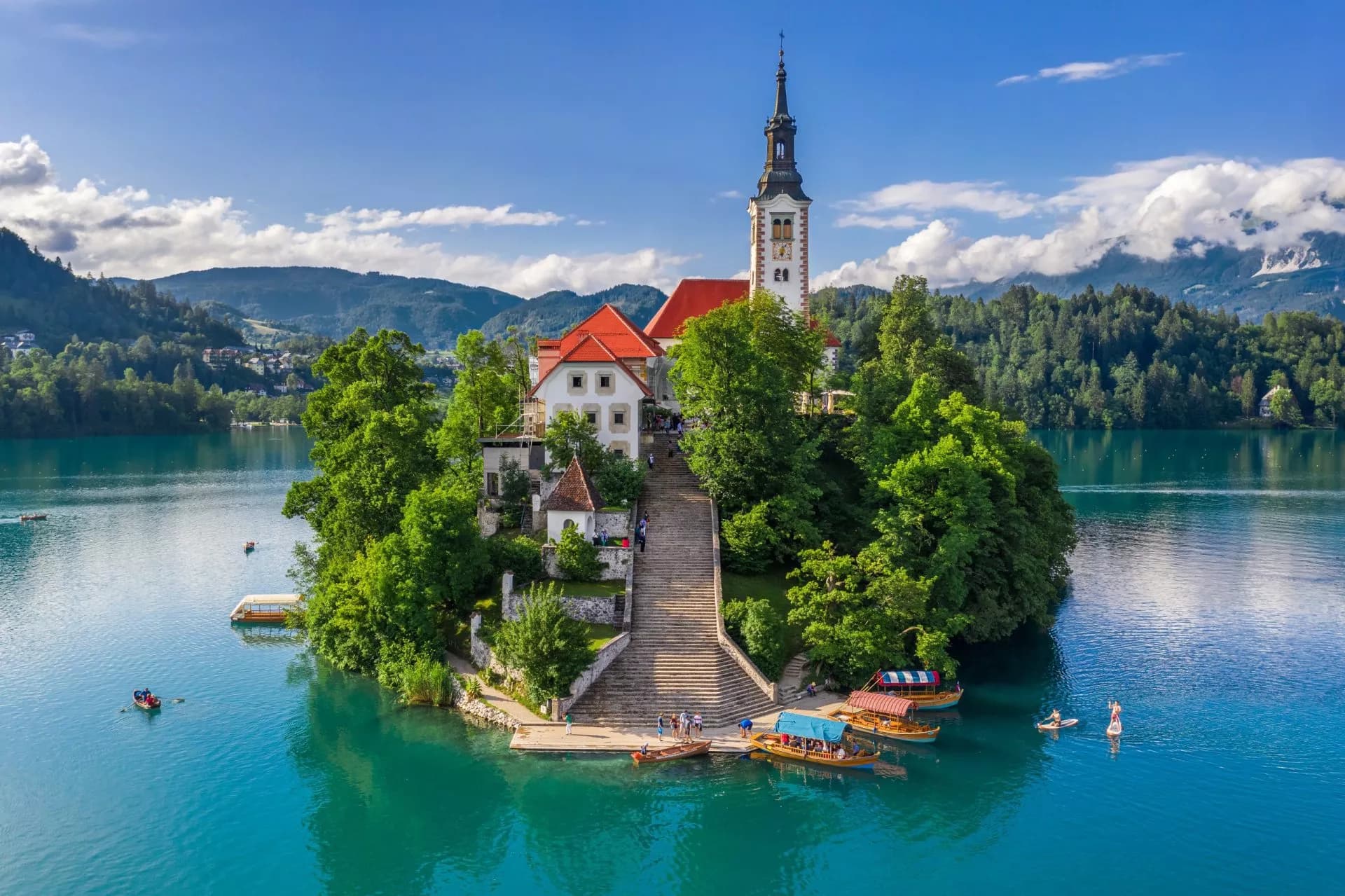 Church on Bled Island in turquoise Lake Bled, Slovenia, with boats and forested mountains.