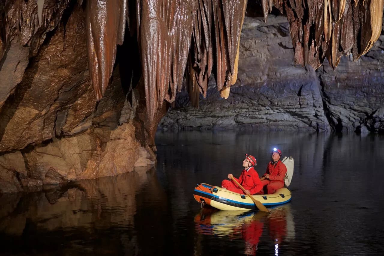 Cave exploration with two people in a raft under large brown stalactites over dark water.