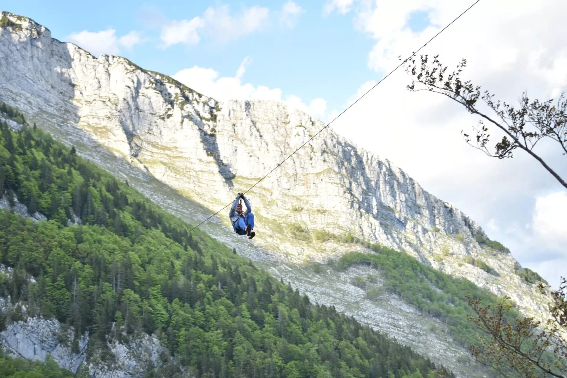 Ziplining over a steep, forested mountain slope with a sheer white rock face under a blue sky in Bovec.