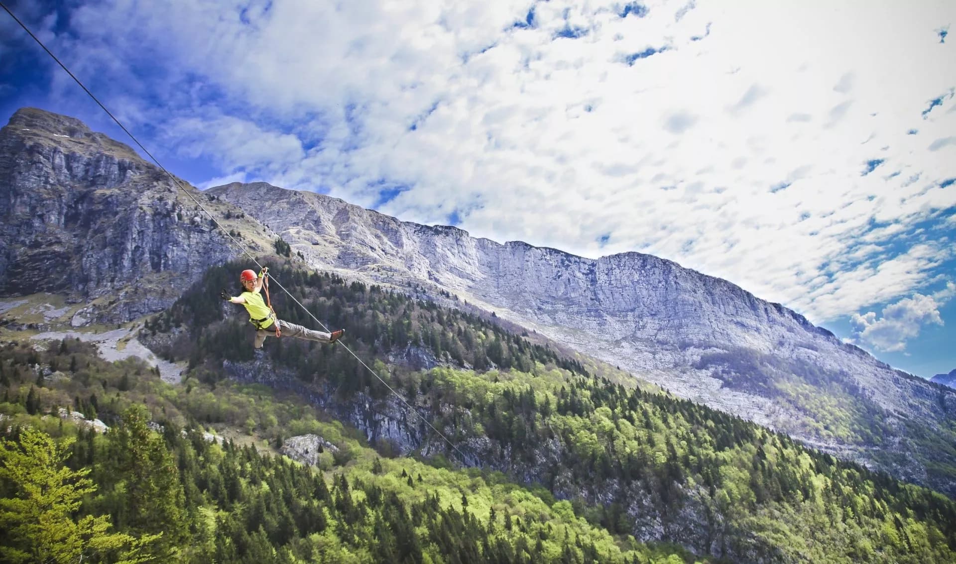 Zip-lining across a deep valley with steep, forested mountains under a cloudy blue sky in Soca Valley.