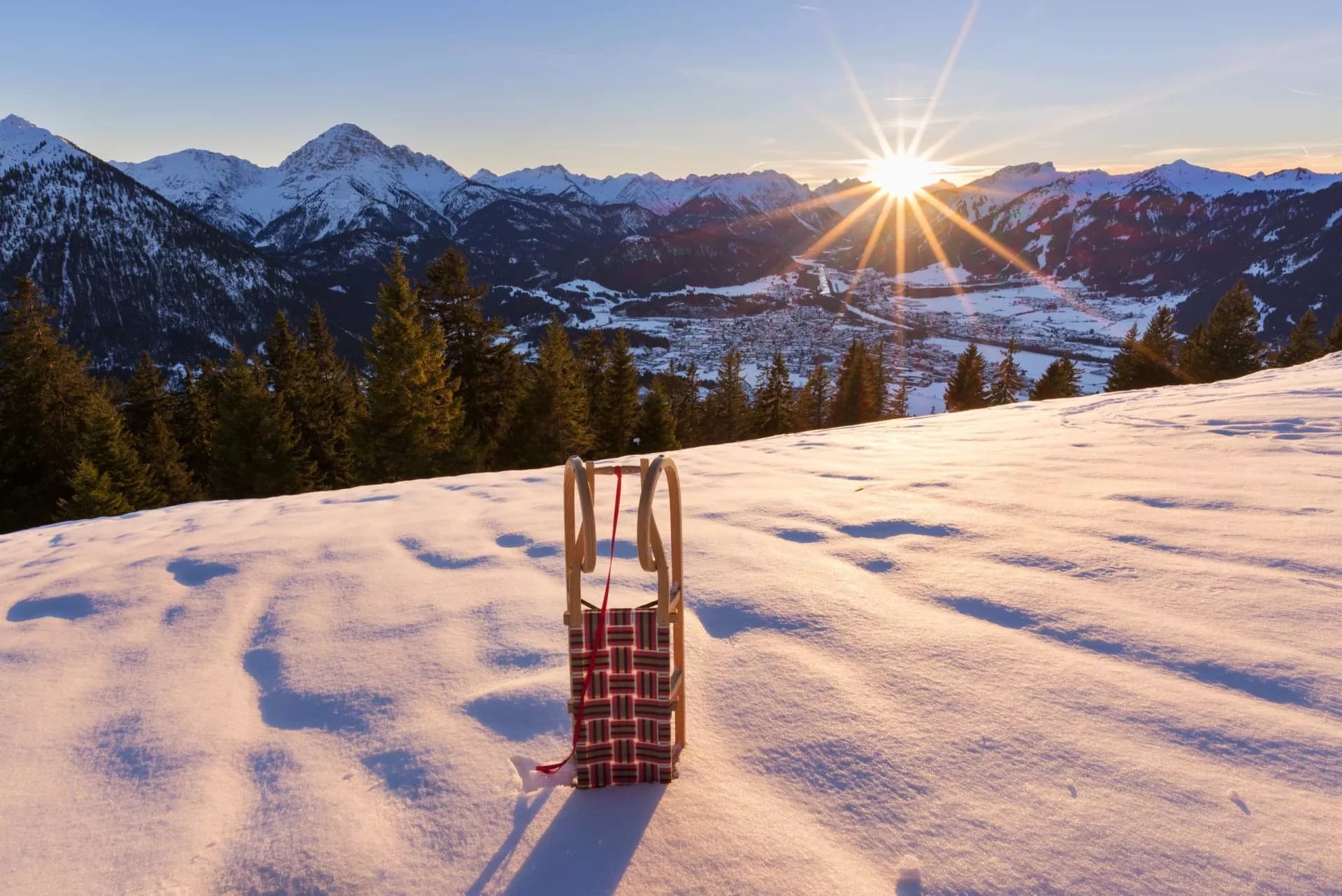 Wooden sled on snowy slope overlooking an alpine town and sunlit mountains in Slovenia.