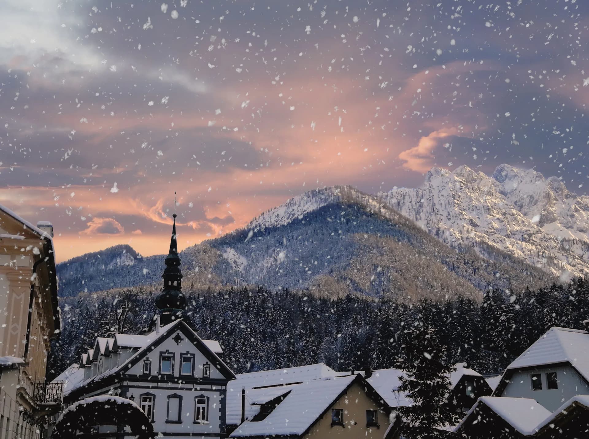 Snow falling over Kranjska Gora village with snow-covered mountains and pink sunset sky.