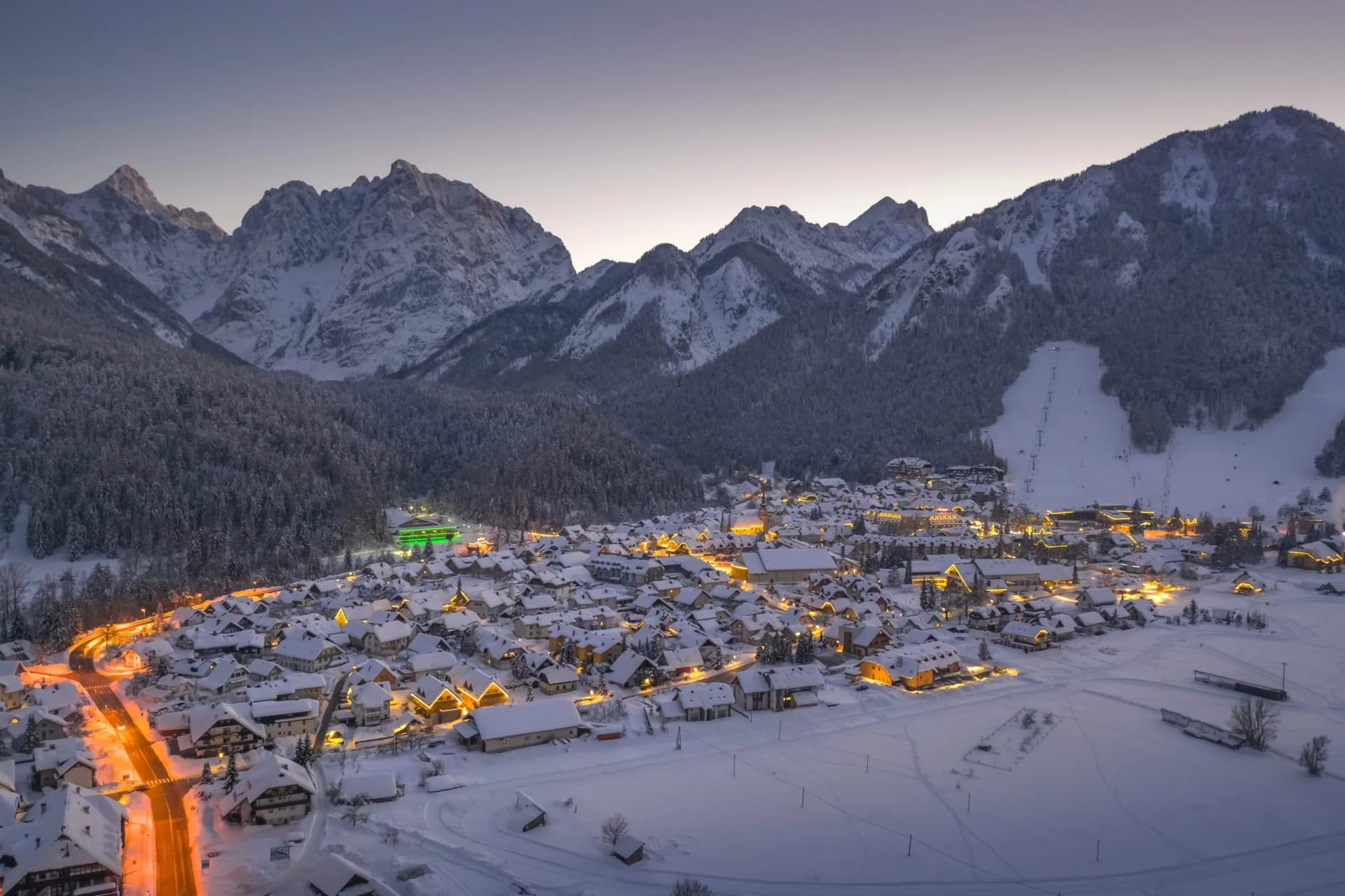 Kranjska Gora village illuminated at dusk in winter with snow-covered houses and mountains.